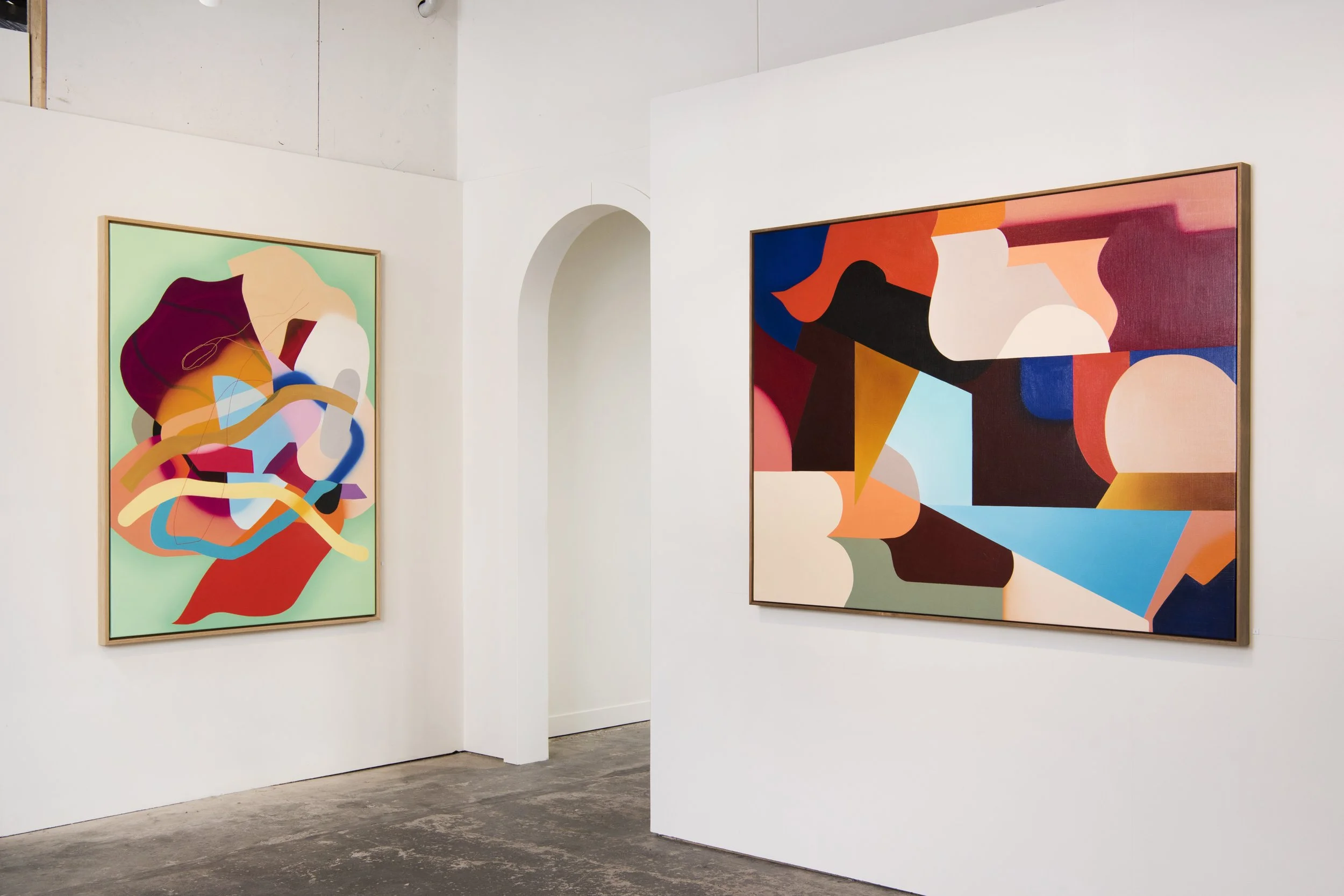 Two colorful abstract paintings on white gallery walls, one on the left with swirling shapes and vibrant colors, the other on the right with geometric blocks in bold hues.