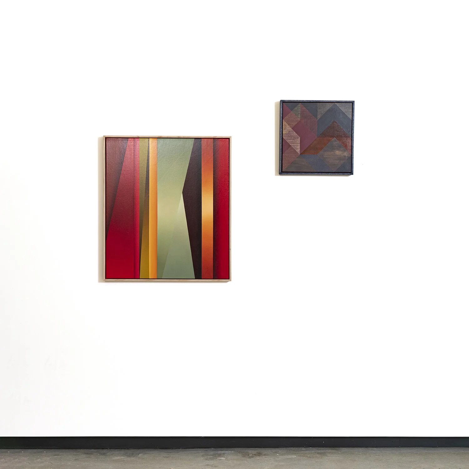 Two abstract geometric paintings on a white gallery wall, one larger with vertical stripes in warm colors, and one smaller with diamond shapes in darker hues.
