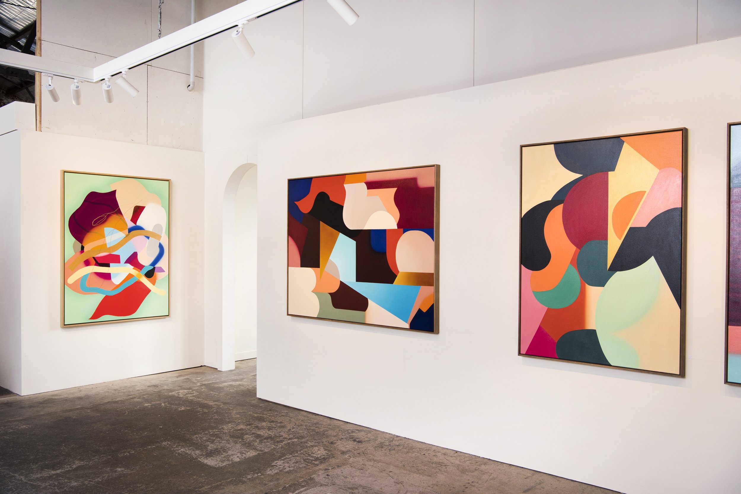 Three abstract colorful paintings displayed on a white gallery wall with a concrete floor.