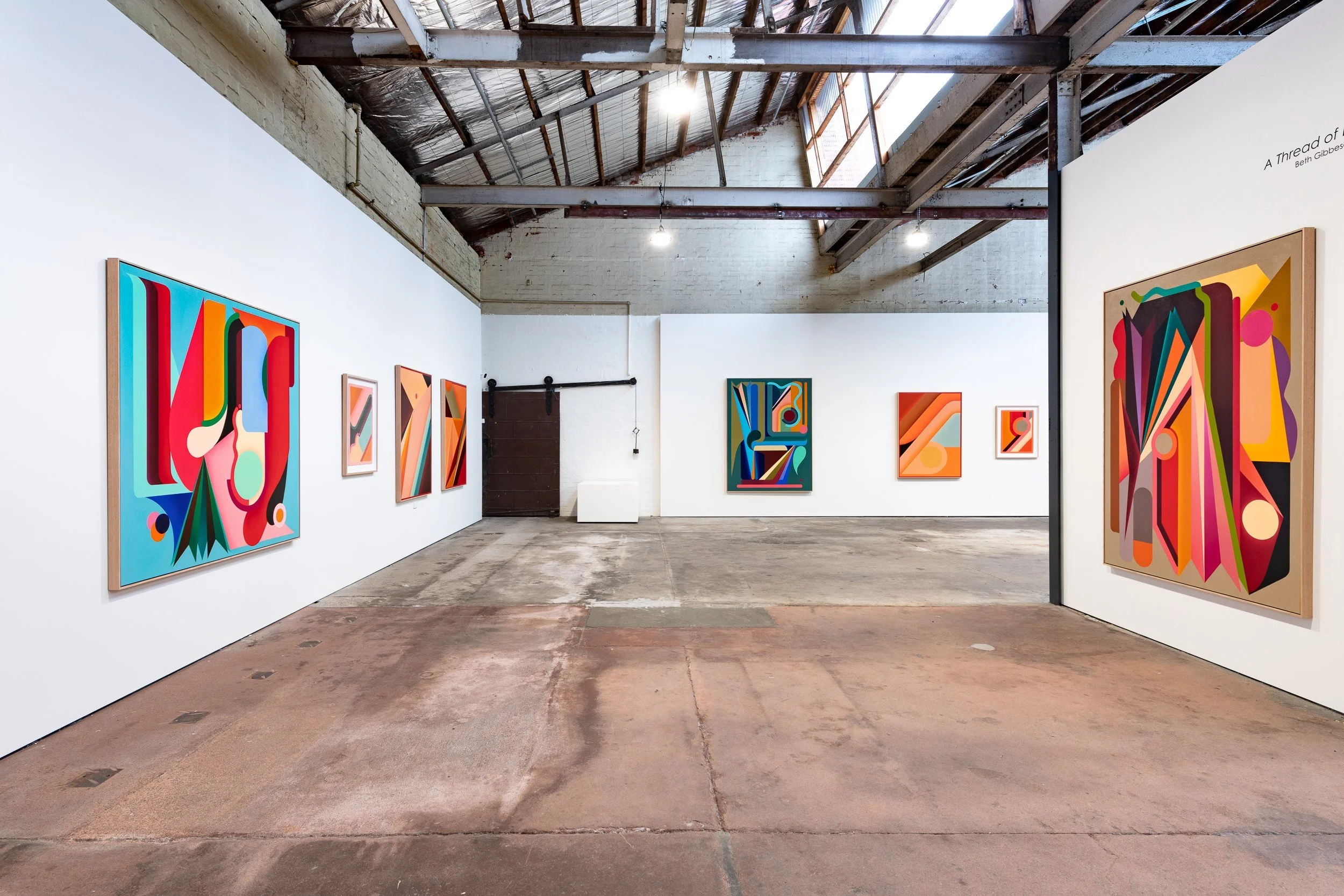 An art gallery with abstract colorful paintings on white walls, visible in an industrial-style space with a high ceiling, exposed beams, and a concrete floor.
