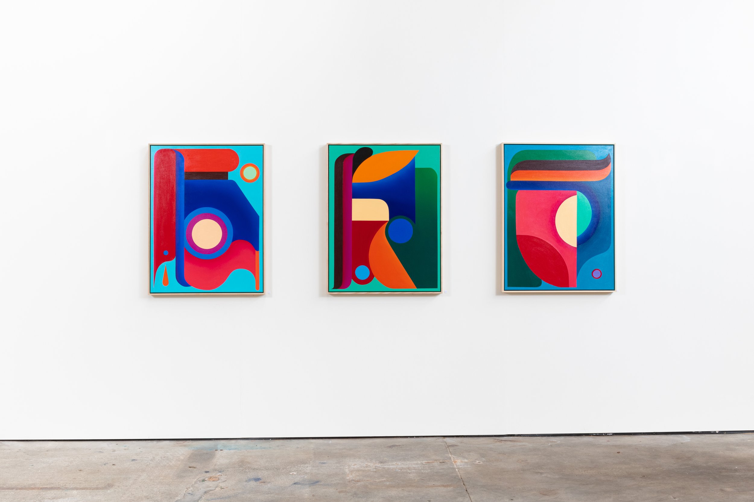 Three colorful abstract paintings in frames hanging on a white gallery wall.