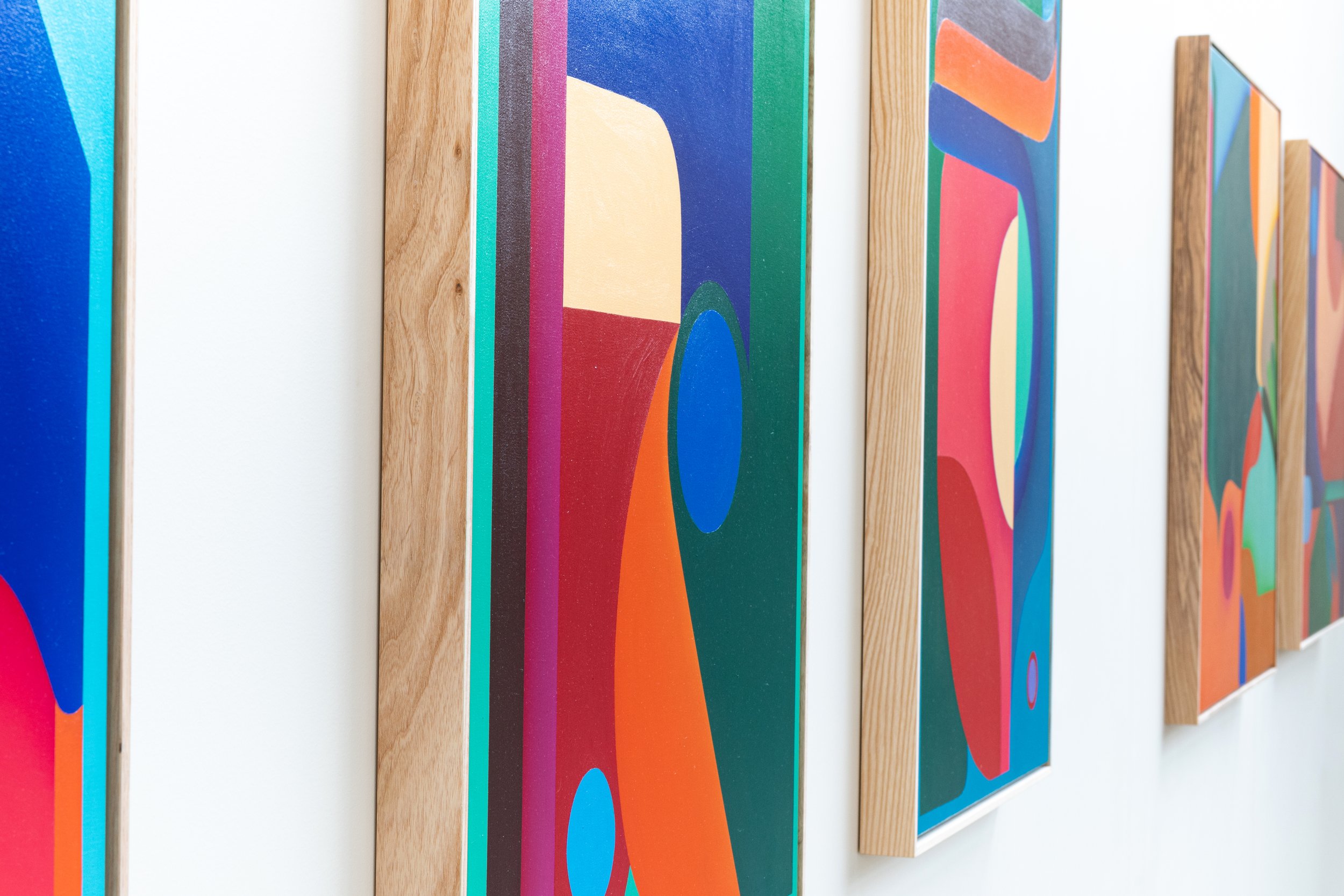 A series of abstract geometric art pieces with vibrant colors and shapes, framed with wood, displayed on a white gallery wall.