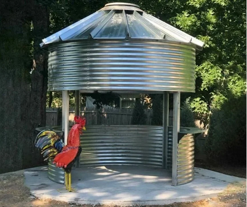 Metal chicken sculpture in front of a small metal well structure with a conical roof, situated outdoors near trees.