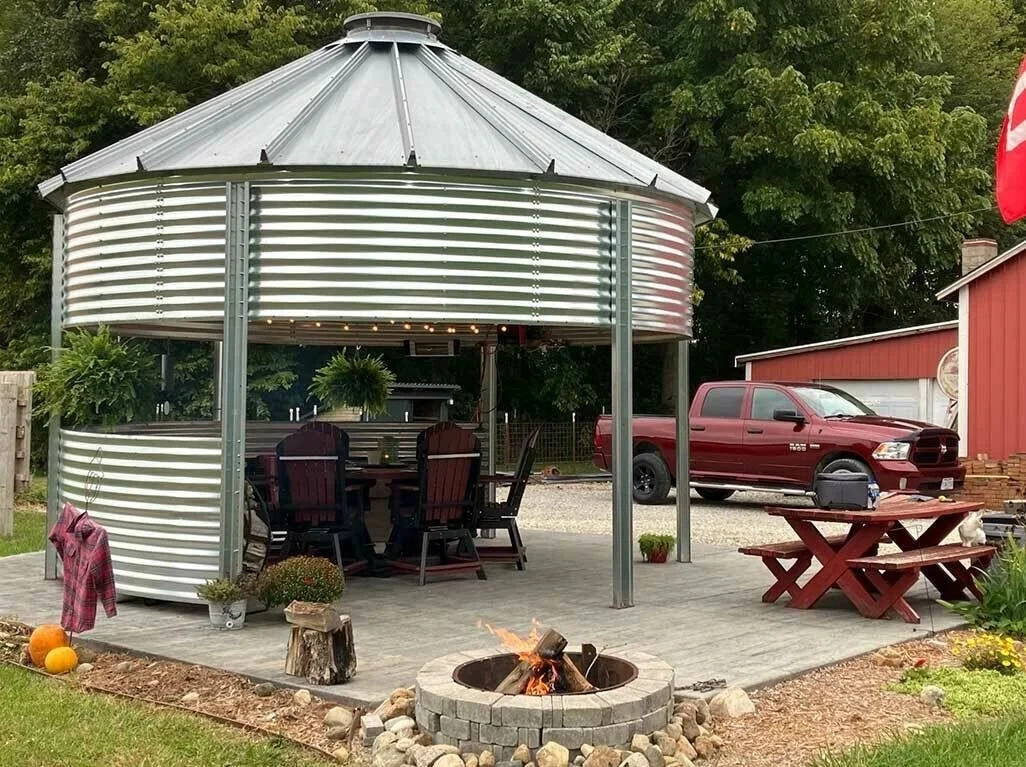 A circular metal pavilion with a conical roof, seating area with a round table and chairs, a fire pit, a red pickup truck, and outdoor decorations in a backyard.
