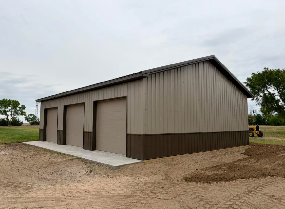 Steel garage with three roll-up doors, located in a rural area with grass, trees, and a tractor in the background.