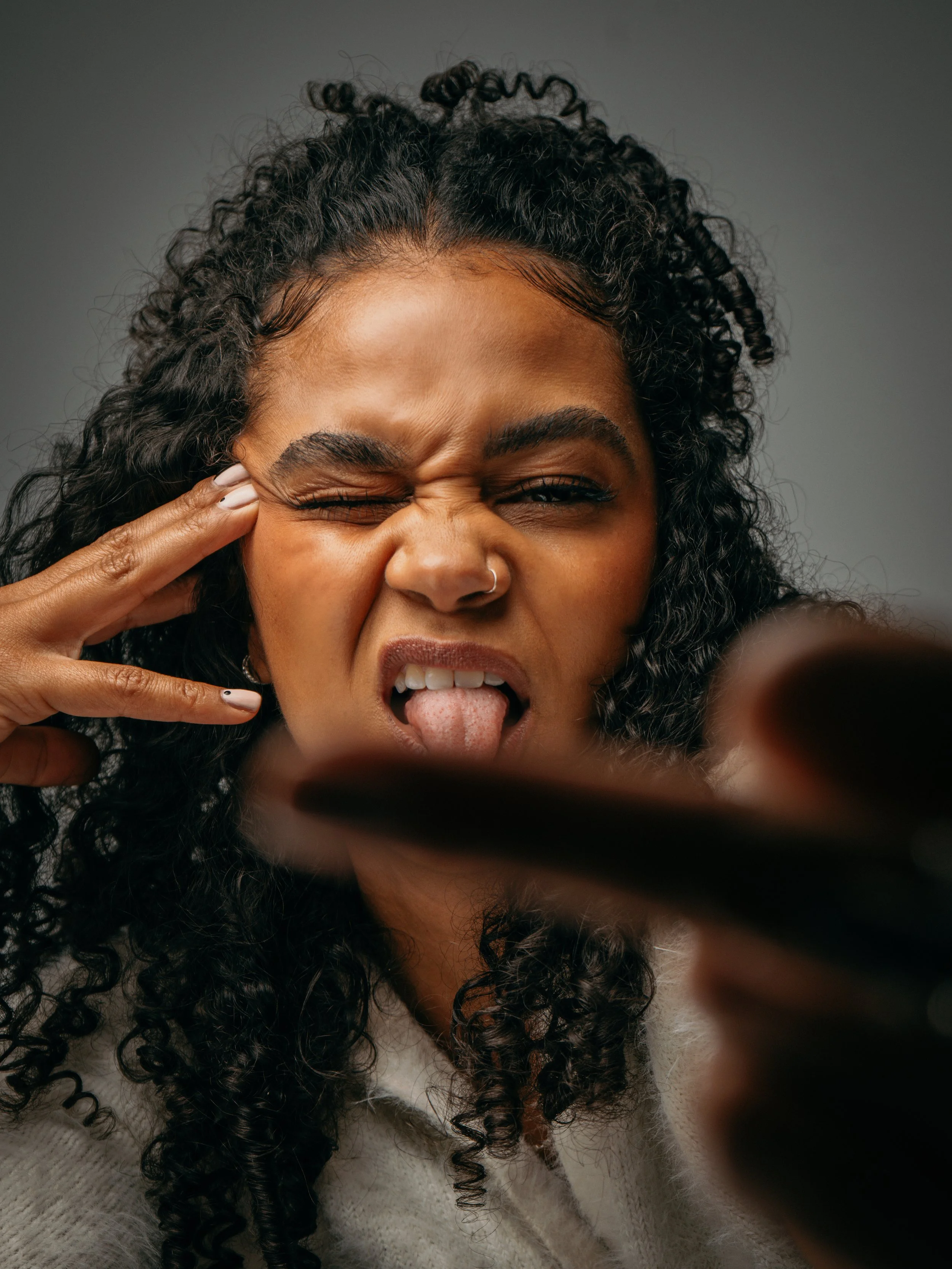 A woman with curly black hair making a face, sticking her tongue out, winking one eye, and tugging at her face with her fingers.