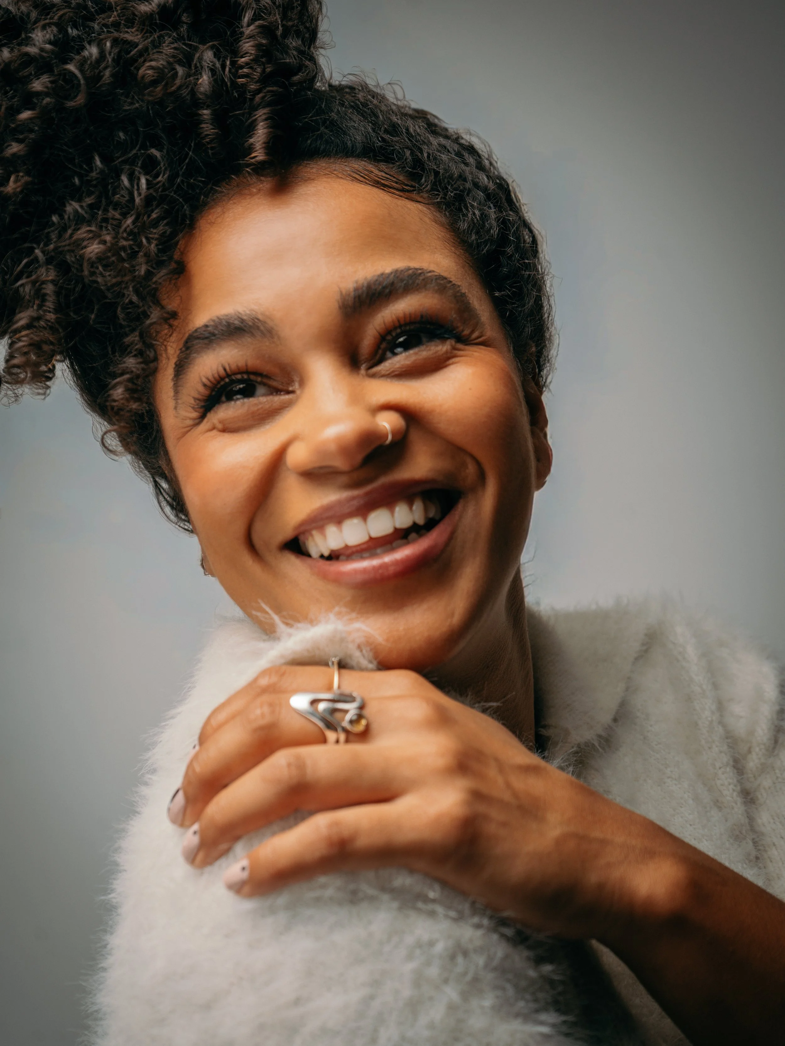 A woman smiling, showing her teeth, with dark curly hair in a textured style, wearing a nose ring, a ring on her finger, and a cozy, light-colored fuzzy sweater.