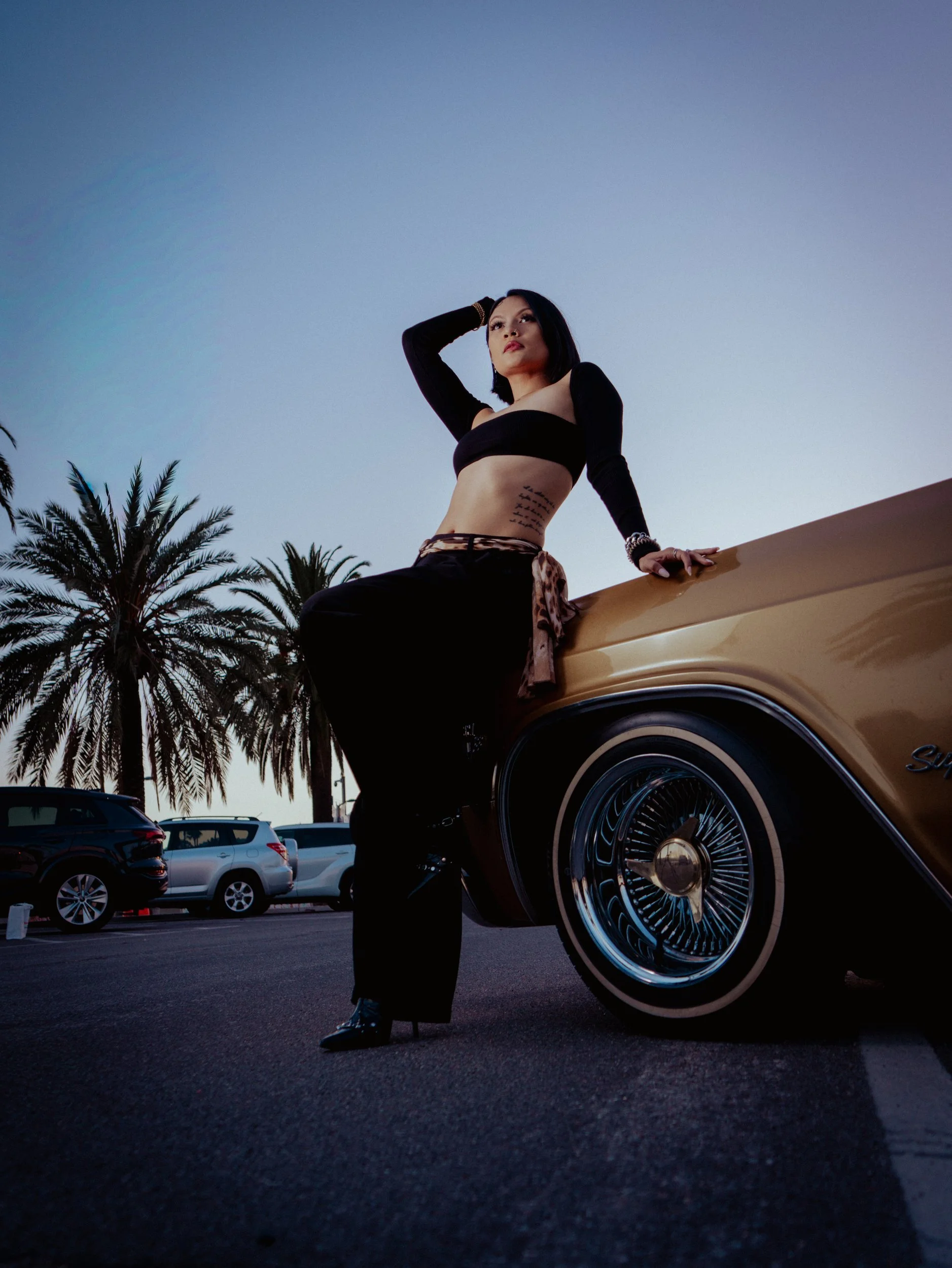 Shaina Rae leaning on a lowrider with palm trees behind her