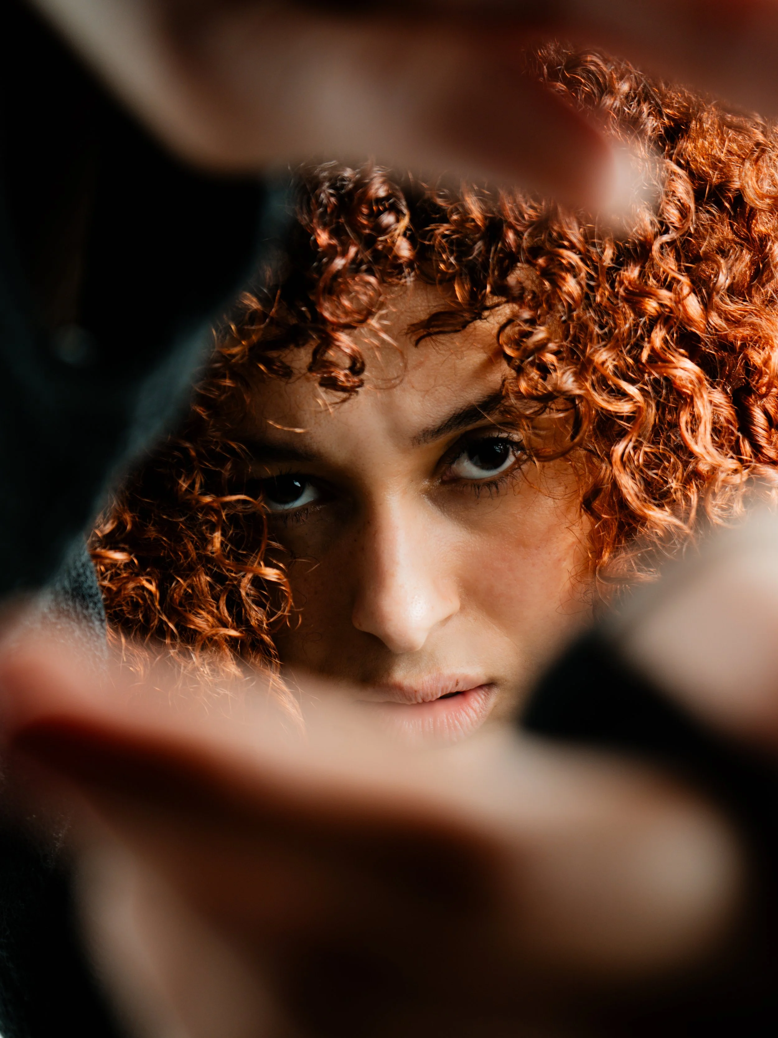 Close-up of a woman with curly red hair peering through a gap, with only her eyes and part of her face visible.