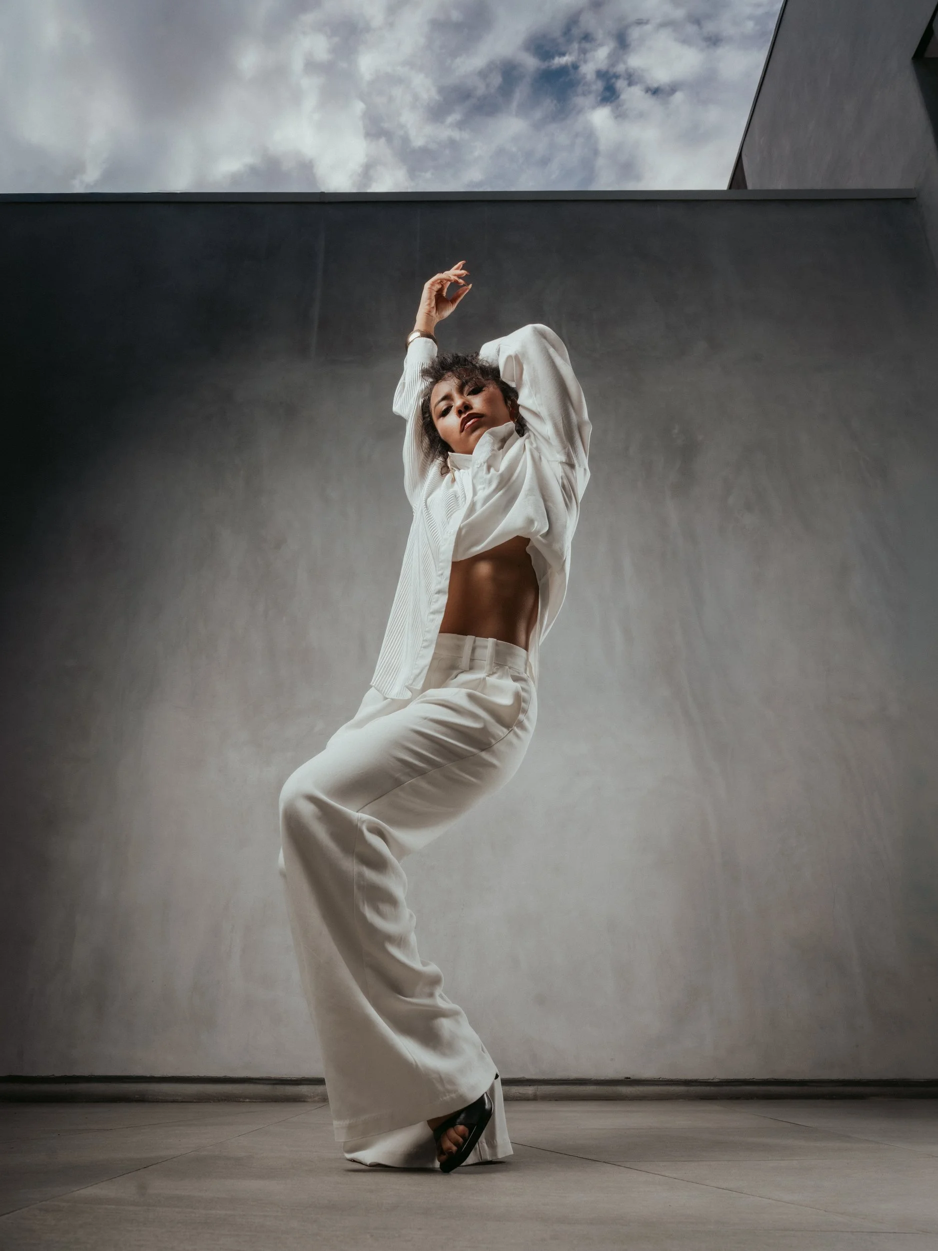 Mirna Moreno in white against a concrete wall and cloudy sky