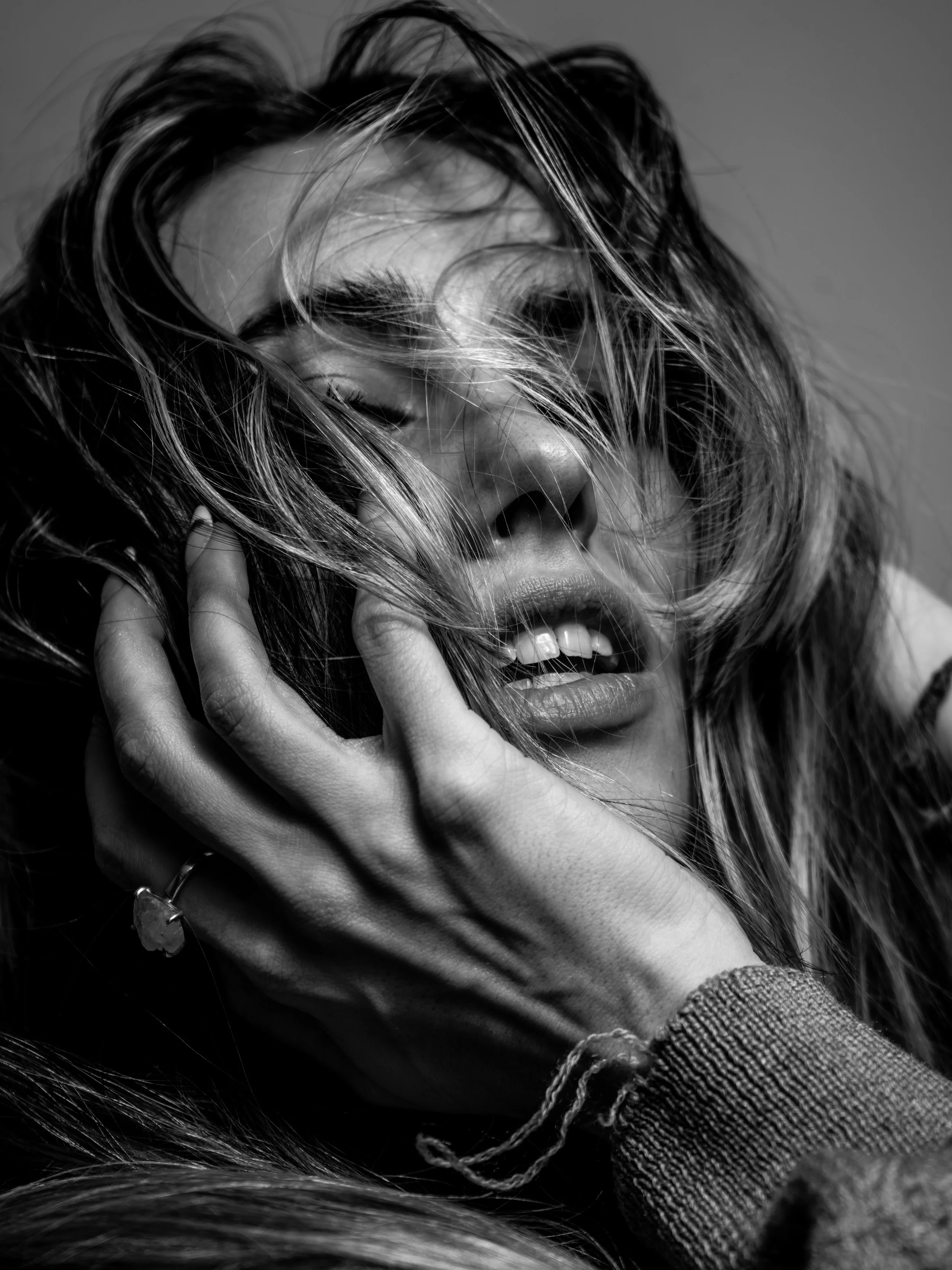A black and white close-up photograph of a person's face with hair covering parts of it. The person has their eyes closed, slightly parted lips, and a hand near their face, touching their hair.

