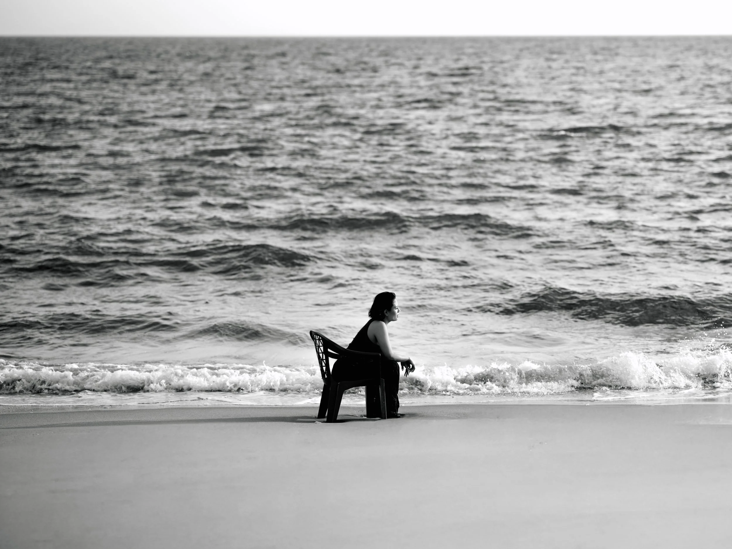 A woman sitting on a chair at the beach looking out at the ocean in black and white.