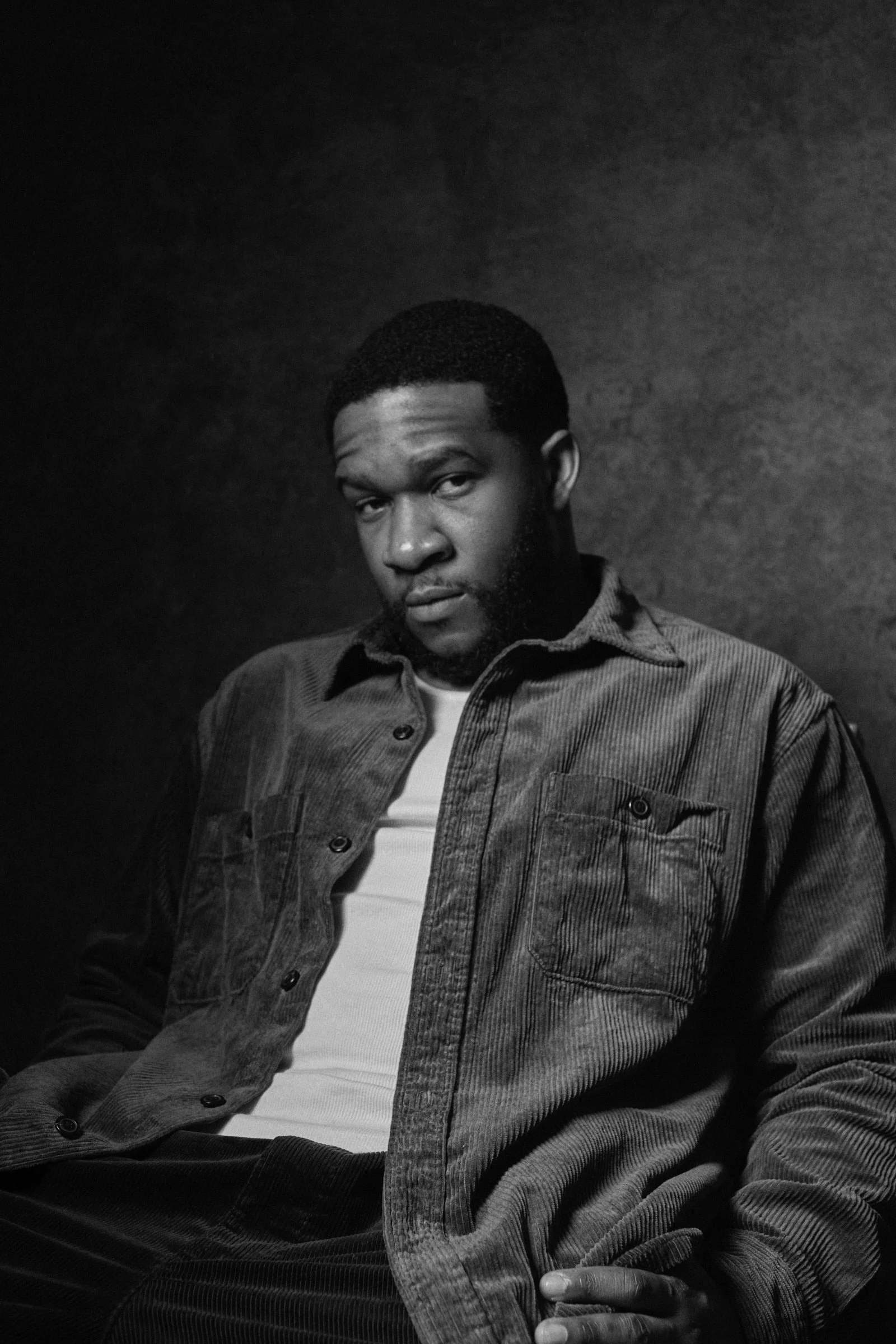Black and white portrait of a young man with short hair and a beard, wearing a denim jacket over a white shirt, sitting against a dark background.