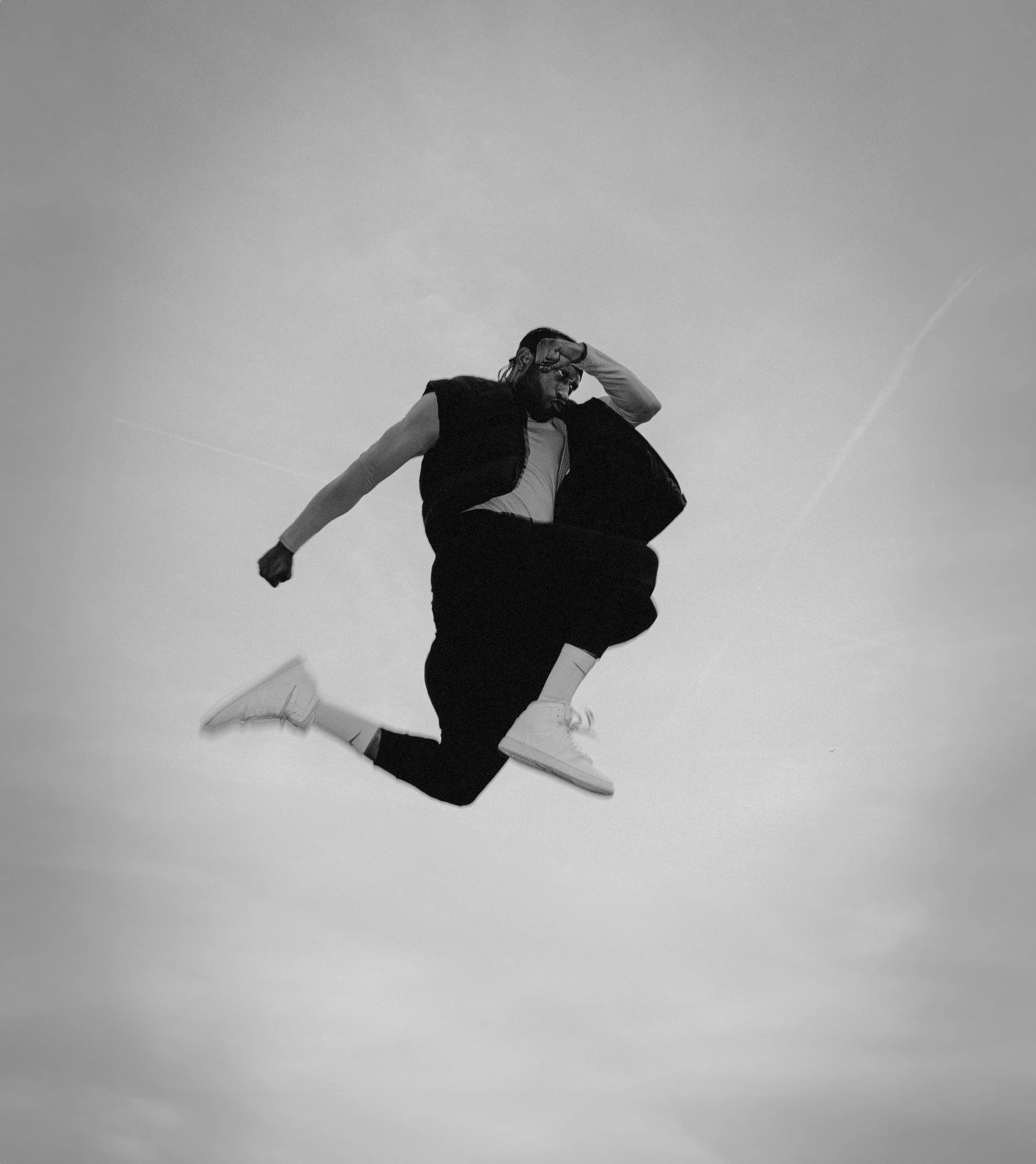 A man in a black sleeveless shirt and white sneakers is mid-air, performing a jump or dance move against a clear sky.