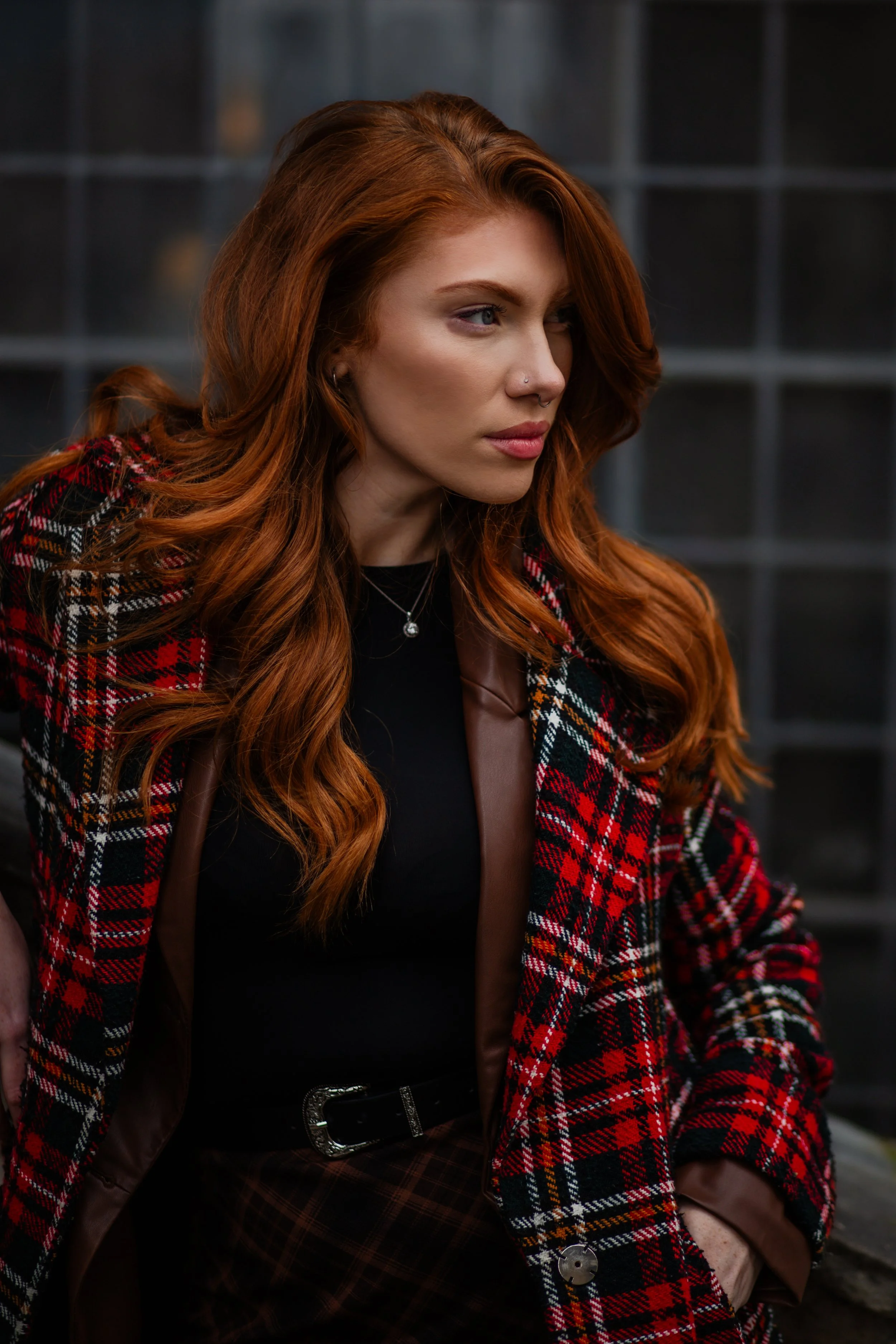 Close-up of a woman with long red hair, wearing a red plaid jacket and black top, seated against a dark tiled background.