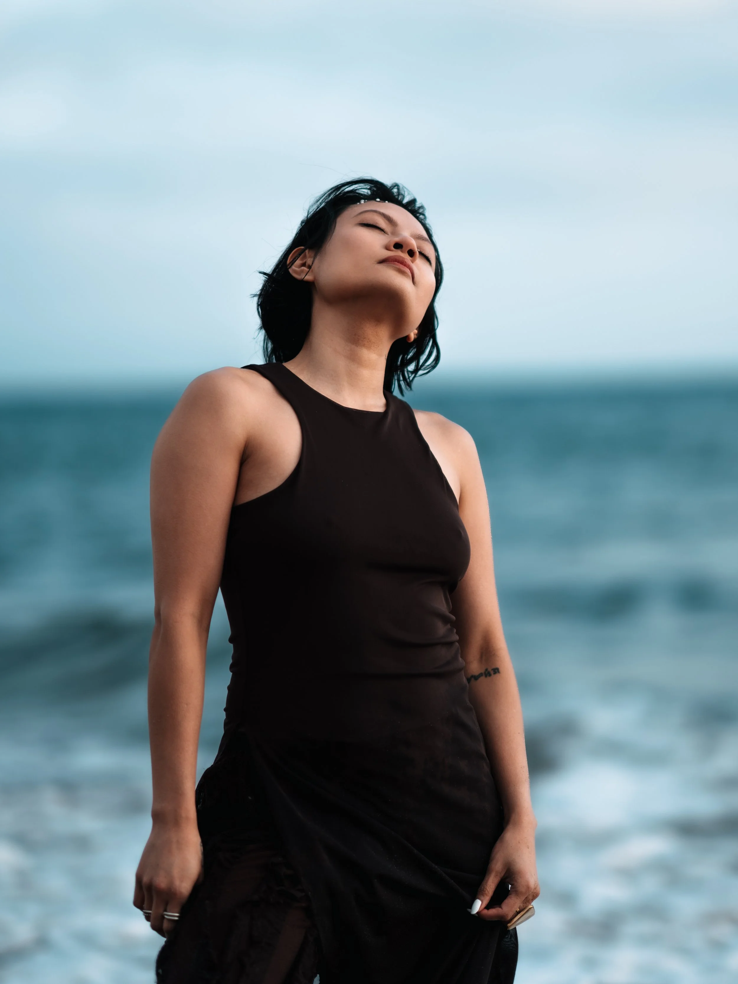 A woman with short black hair standing in front of the ocean with waves, wearing a black sleeveless top and black skirt, eyes closed and smiling slightly.