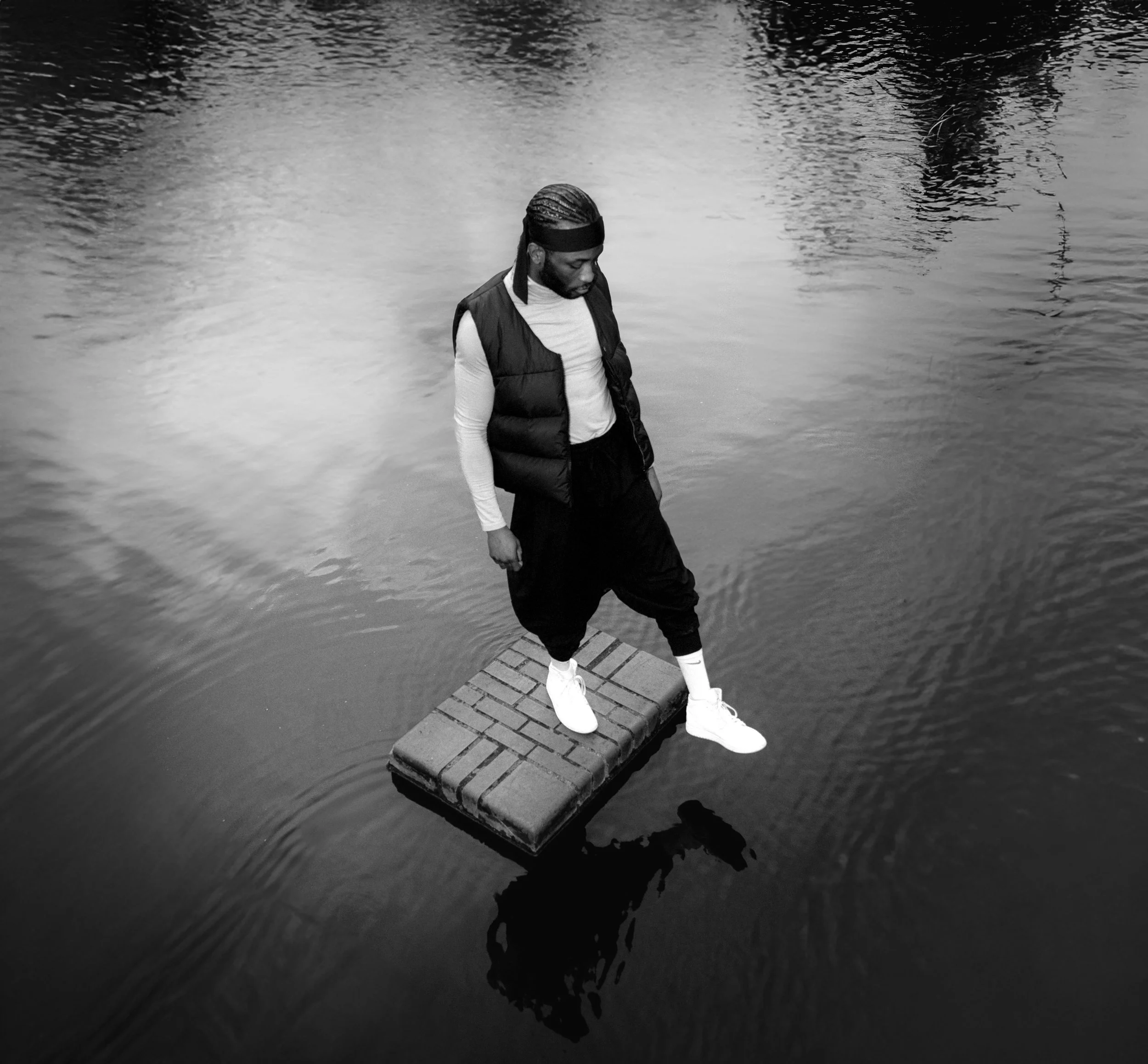 A man with braided hair and wearing a headband, jacket, and sneakers, standing on a wooden dock by a body of water, looking down with his hands in his pockets.