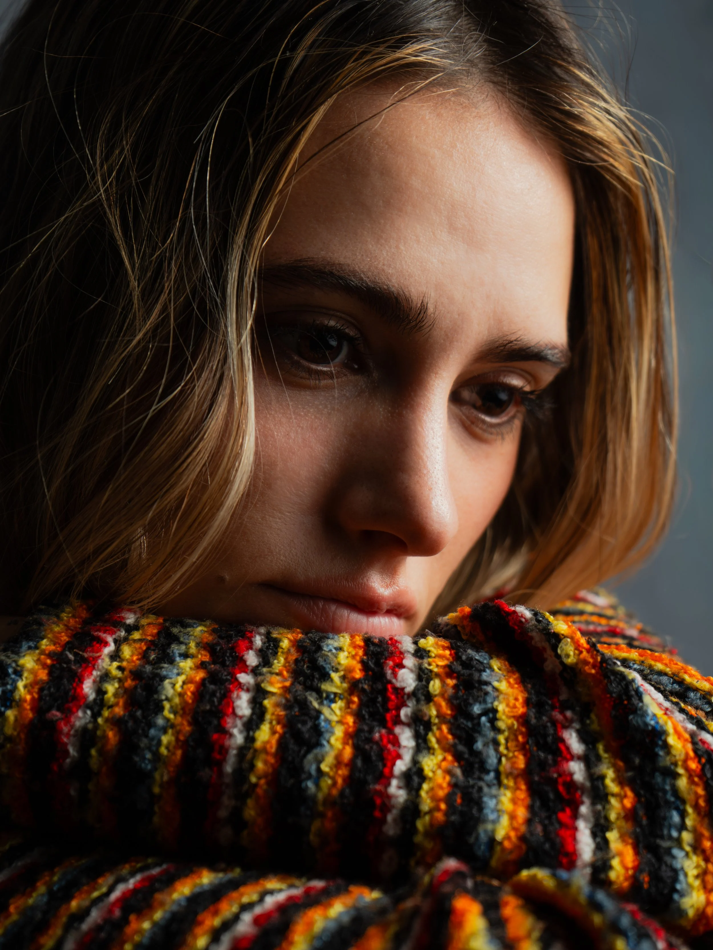 Close-up of a woman's face with wavy brown hair and a colorful, textured sweater, looking downward.