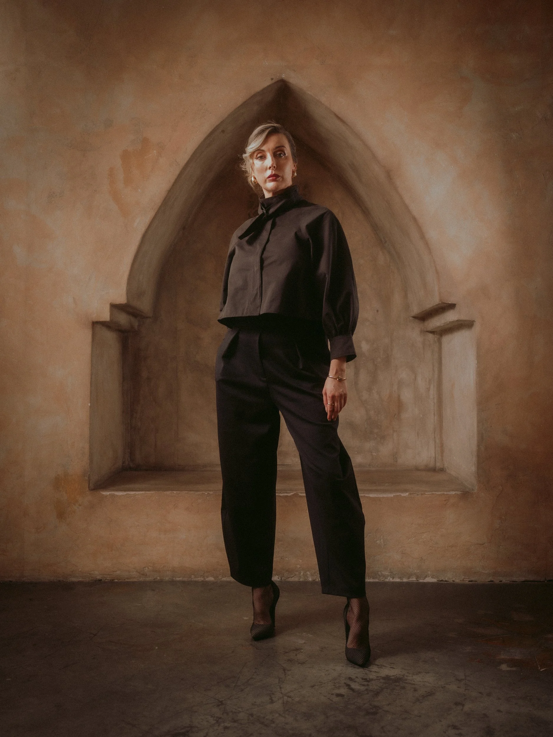 Full-length Milanka Brooks standing in a clay arch in black wide-leg trousers, editorial fashion photography by Osy Ikhile, Los Angeles