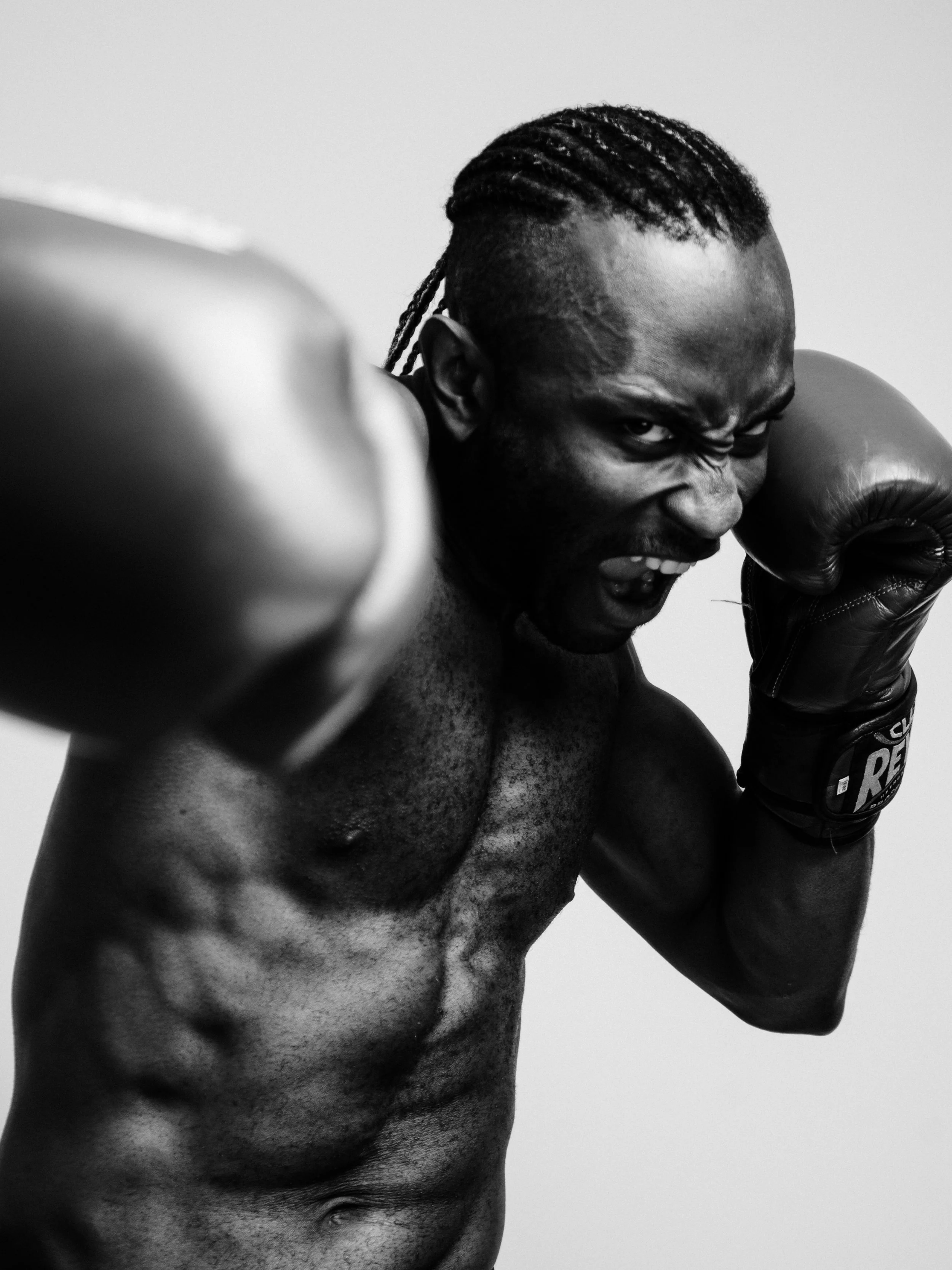 A black and white image of a muscular man with dreadlocks wearing boxing gloves, in a fighting stance with a fierce expression.