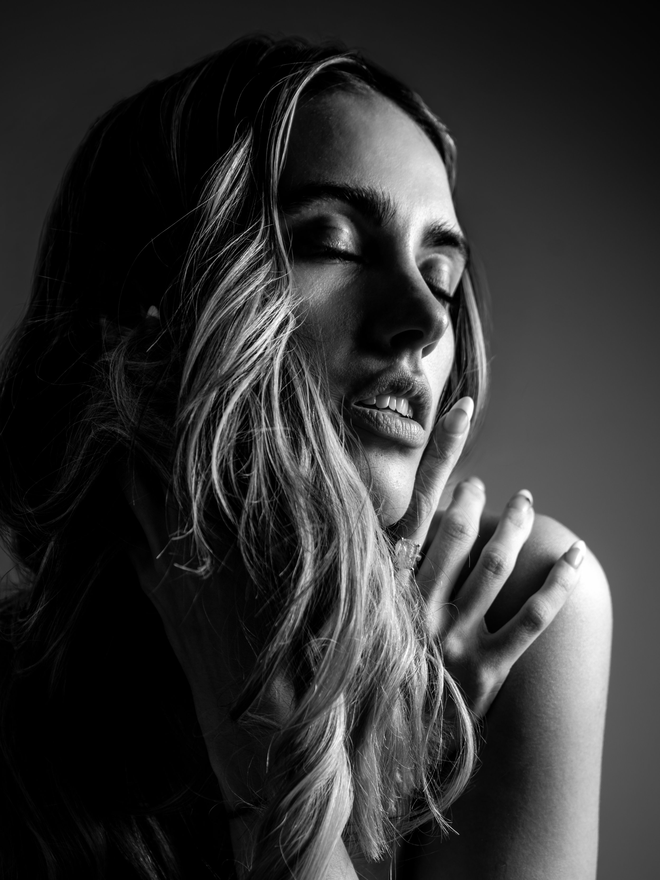 Black and white portrait of a woman with long wavy hair, eyes closed, touching her shoulder with her hand, in a contemplative pose.