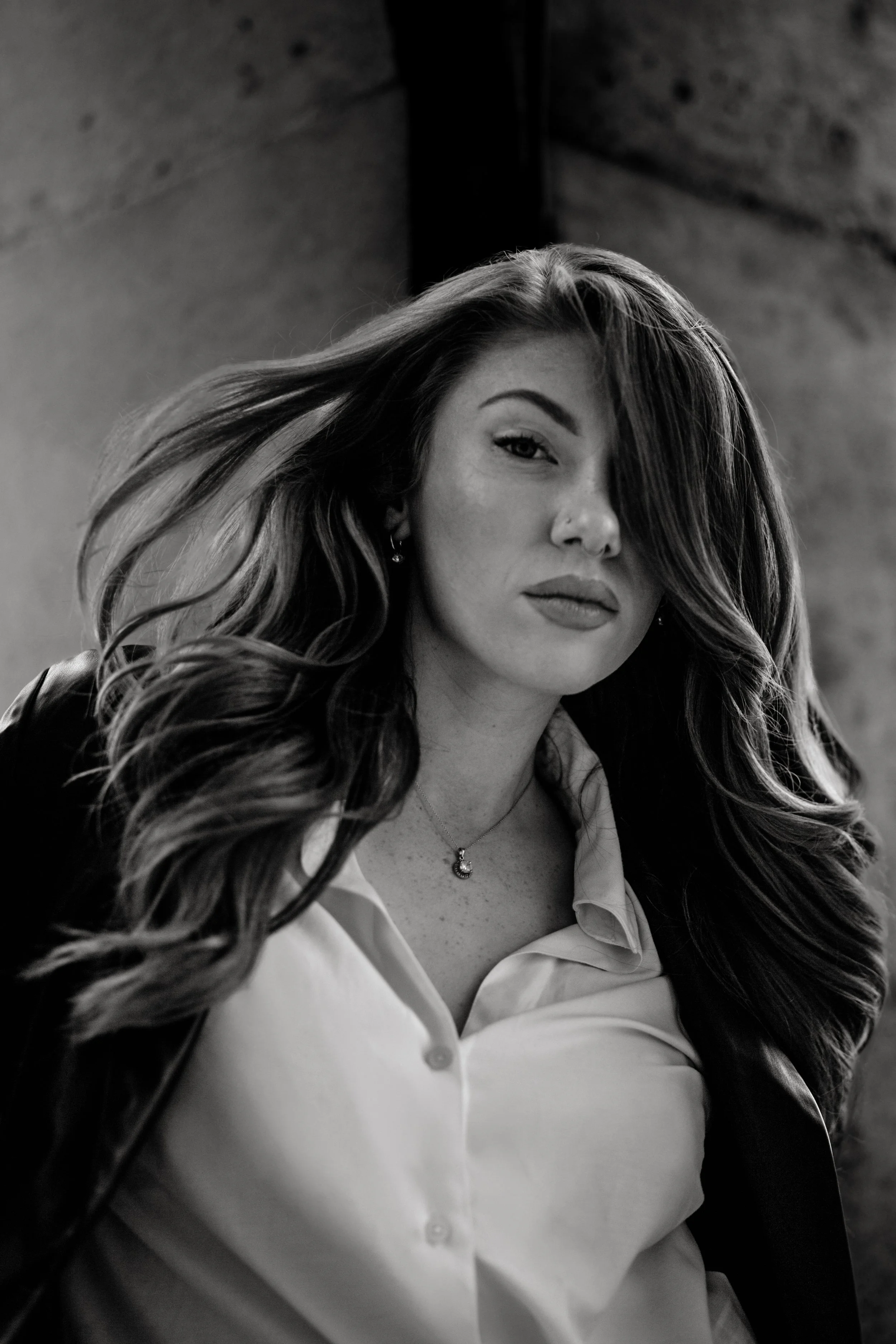 Black and white photo of a woman with wavy hair, wearing a white collared shirt, and a necklace, giving a confident look.