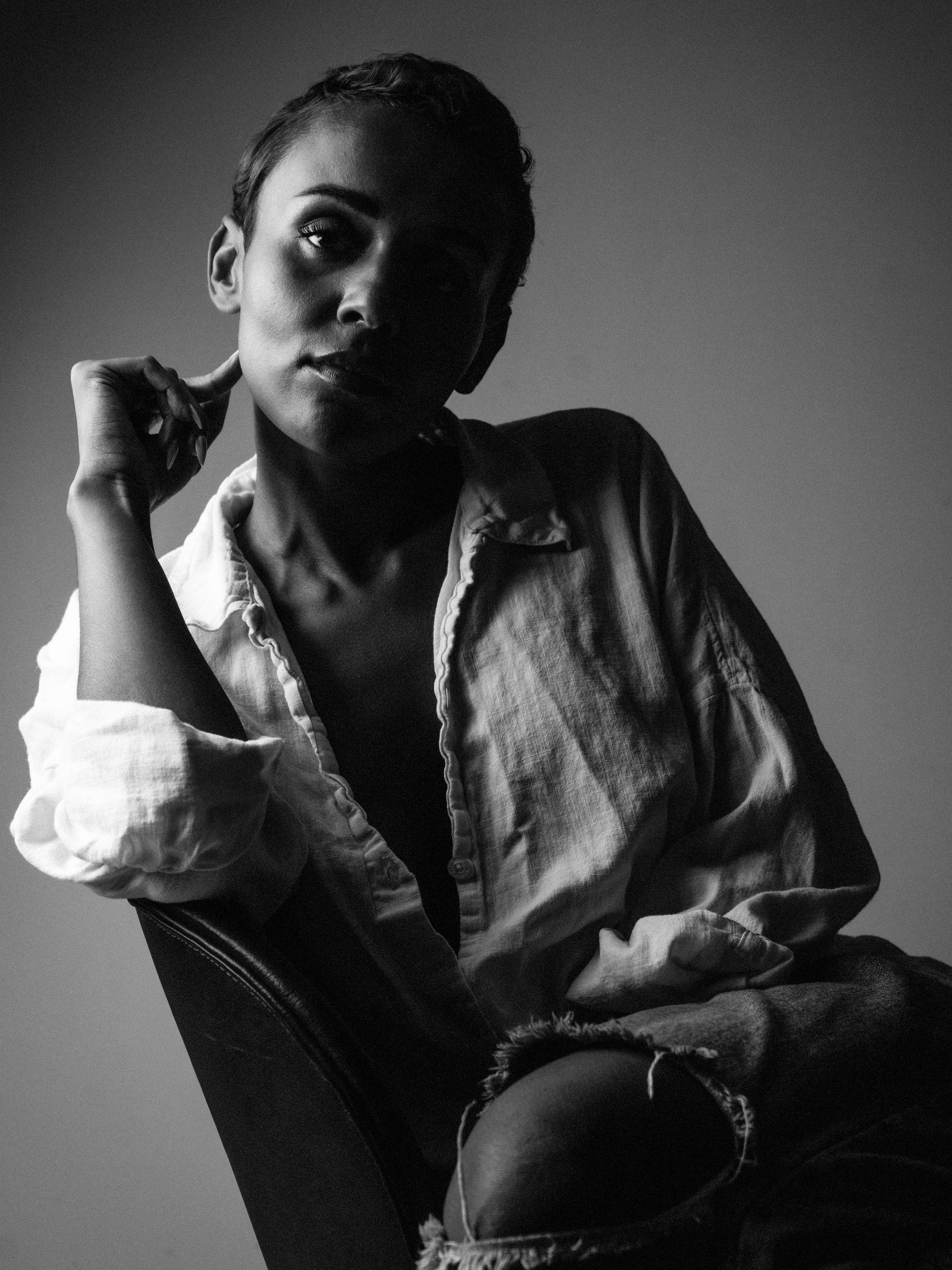 A black and white photograph of a woman with short hair, sitting sideways on a chair, in a thoughtful pose with her left hand touching her neck and her gaze directed upward. She is wearing a slightly unbuttoned shirt with rolled-up sleeves and ripped