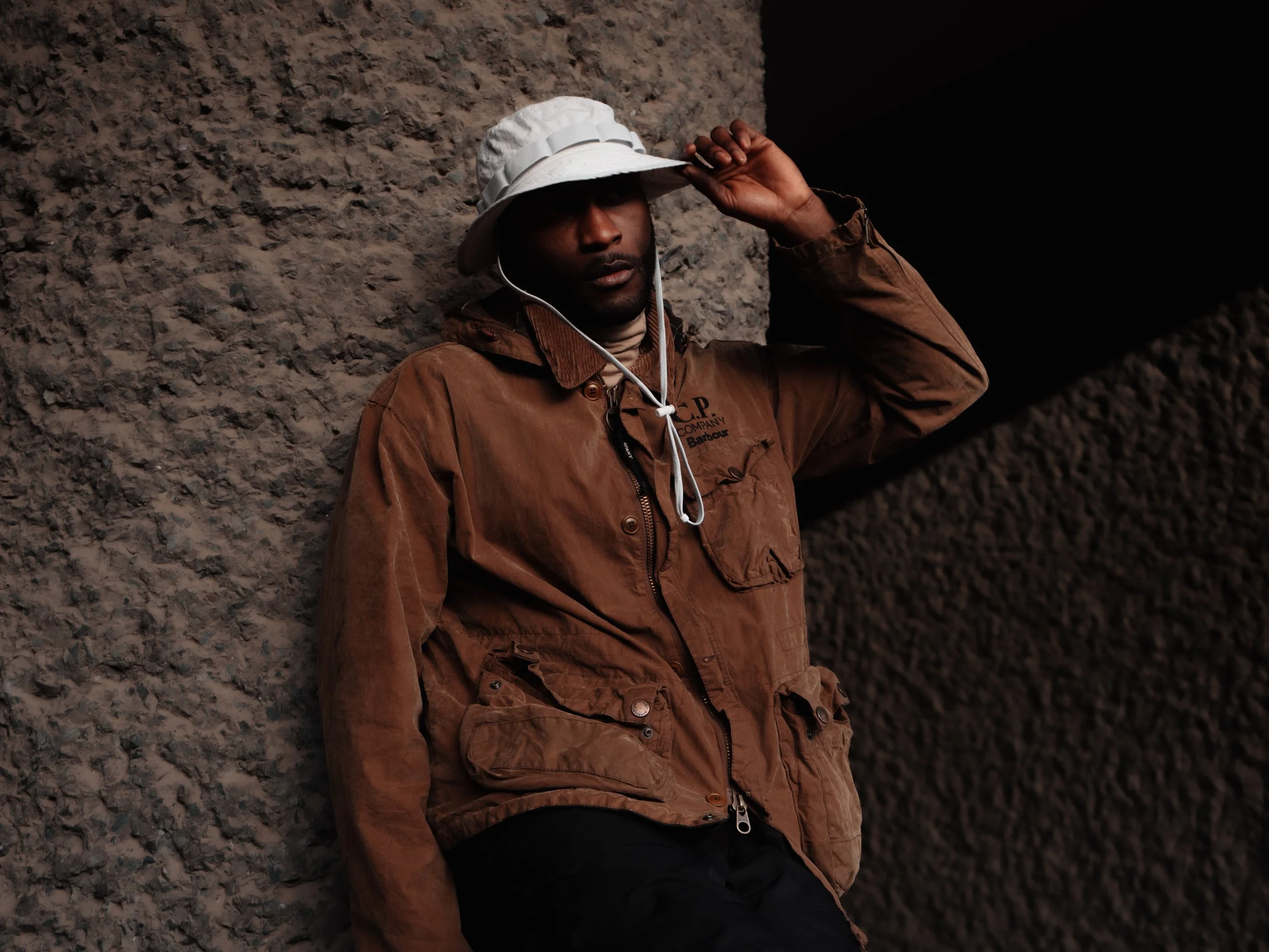 A man wearing a brown jacket and a white bucket hat, adjusting the hat with one hand, standing against a rocky background.