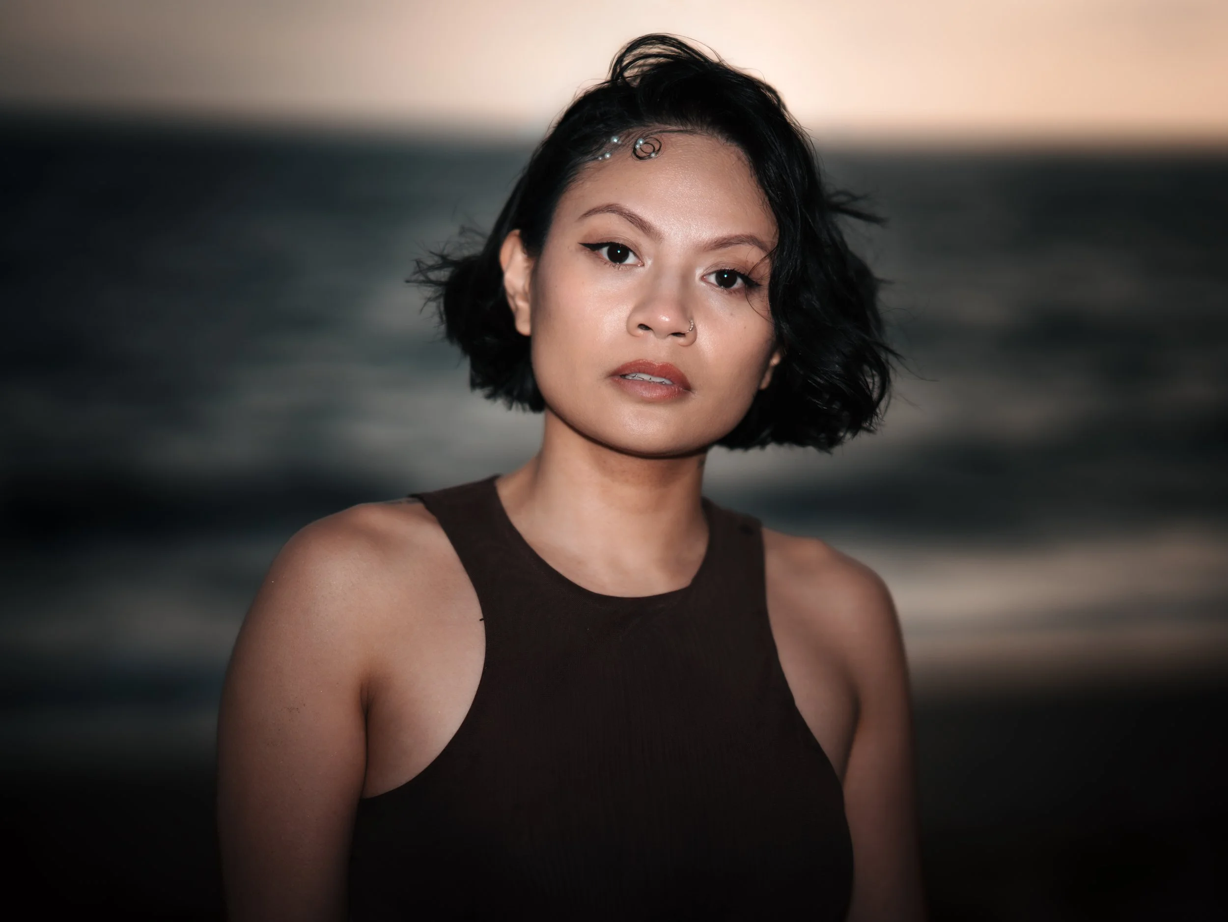 A young woman with short black hair and winged eyeliner, wearing a sleeveless black top, standing outdoors at sunset with the ocean and a partly cloudy sky in the background.