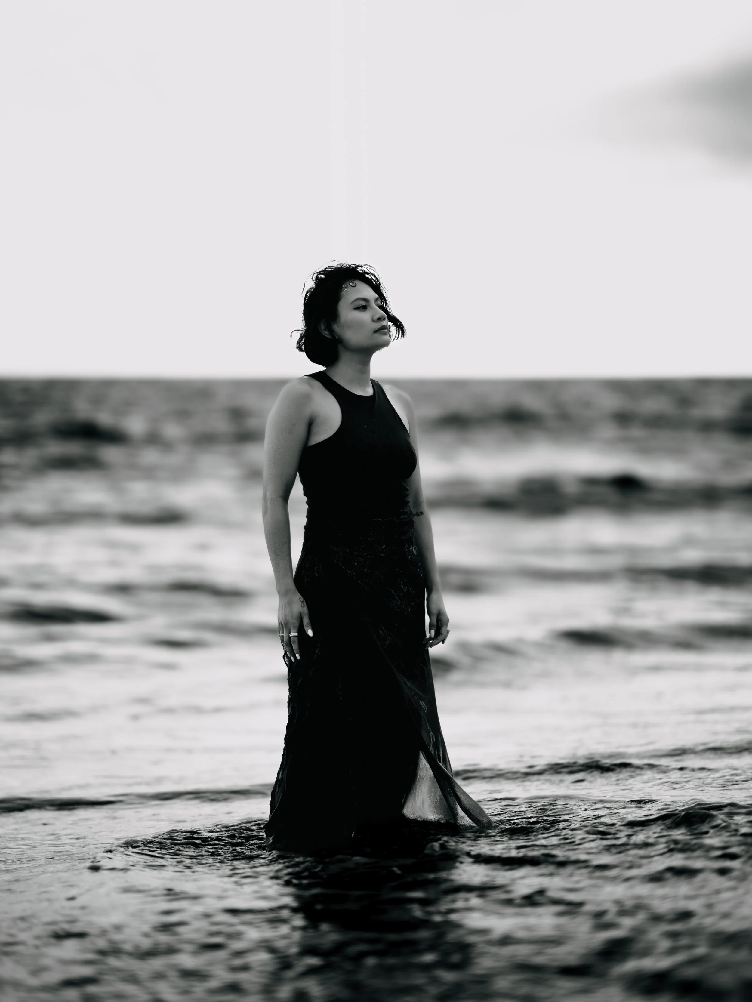 A woman standing in shallow ocean water on a beach, wearing a black dress, with wind-blown hair, looking thoughtfully into the distance.