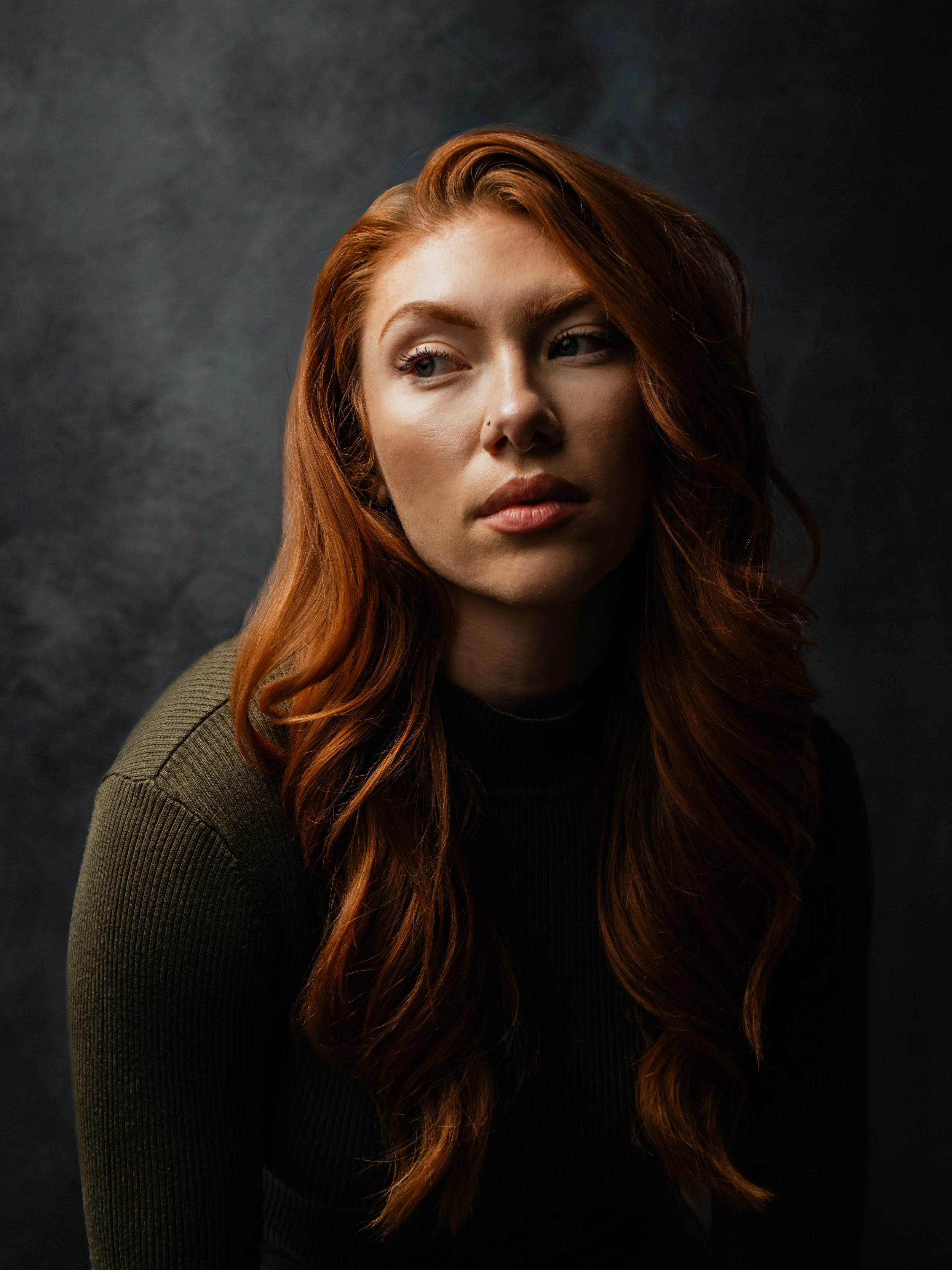 A woman with long red hair looking to her right in a dark setting.