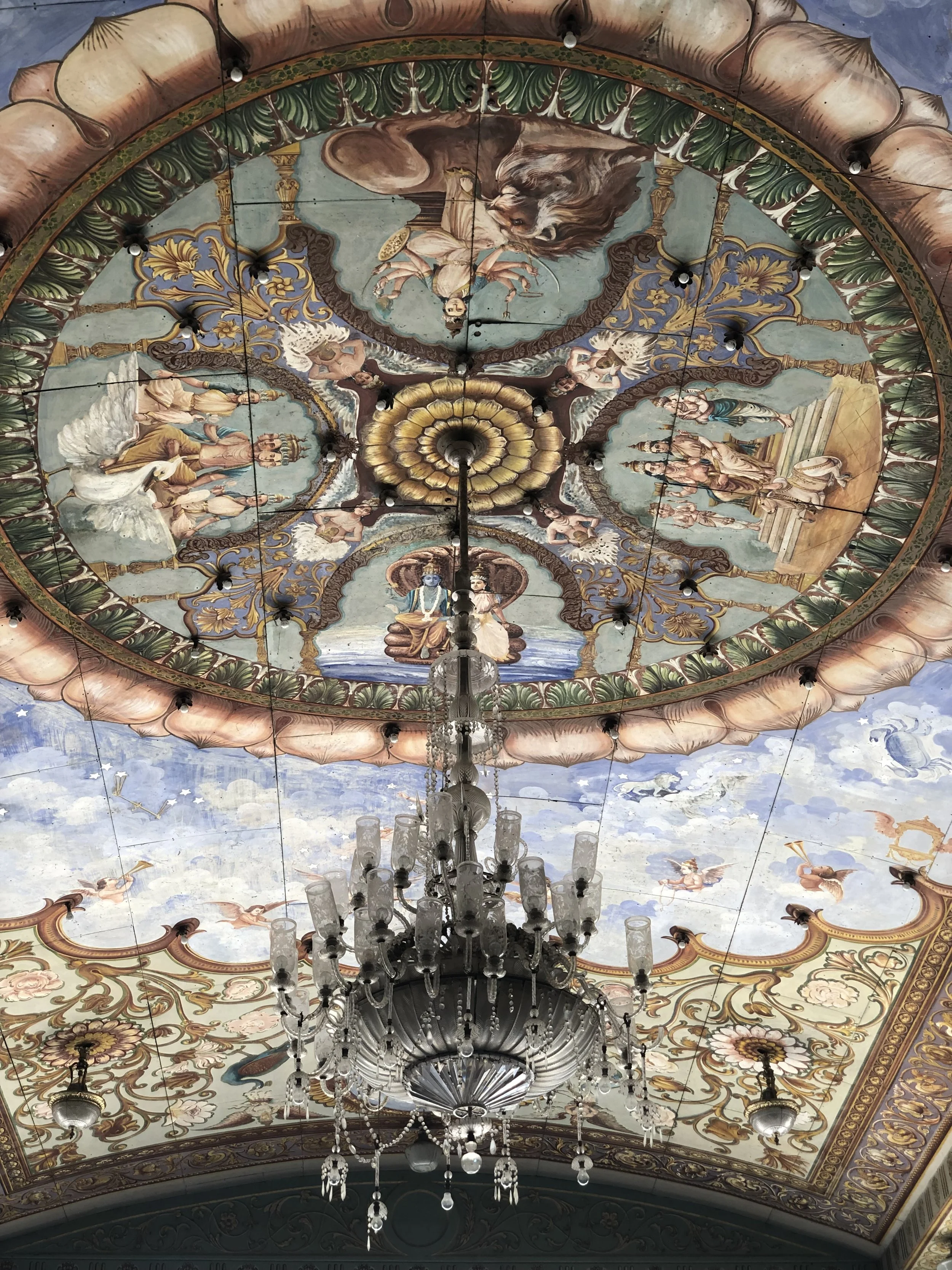 An ornate ceiling with painted murals depicting mythological and historical scenes, adorned with a large crystal chandelier hanging from the center.