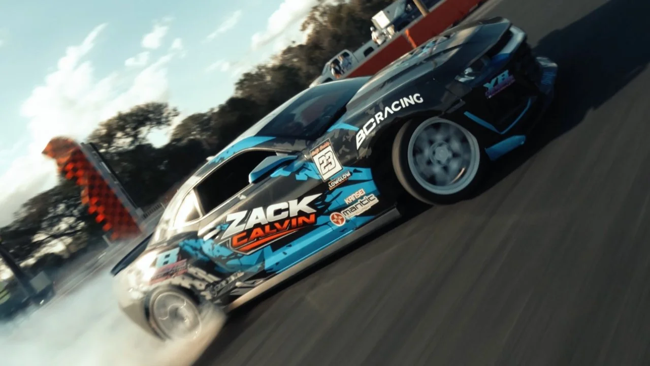 Drone footage of a race car on the track with colorful graphics and sponsor logos, moving at high speed and creating tire smoke.