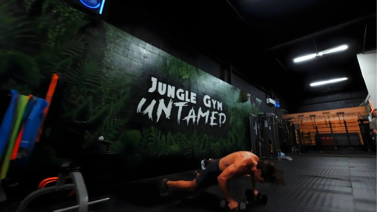 Drone footage of a man is exercising with dumbbells on the floor inside a gym with a jungle-themed wall that reads 'Jungle Gym Untamed'.