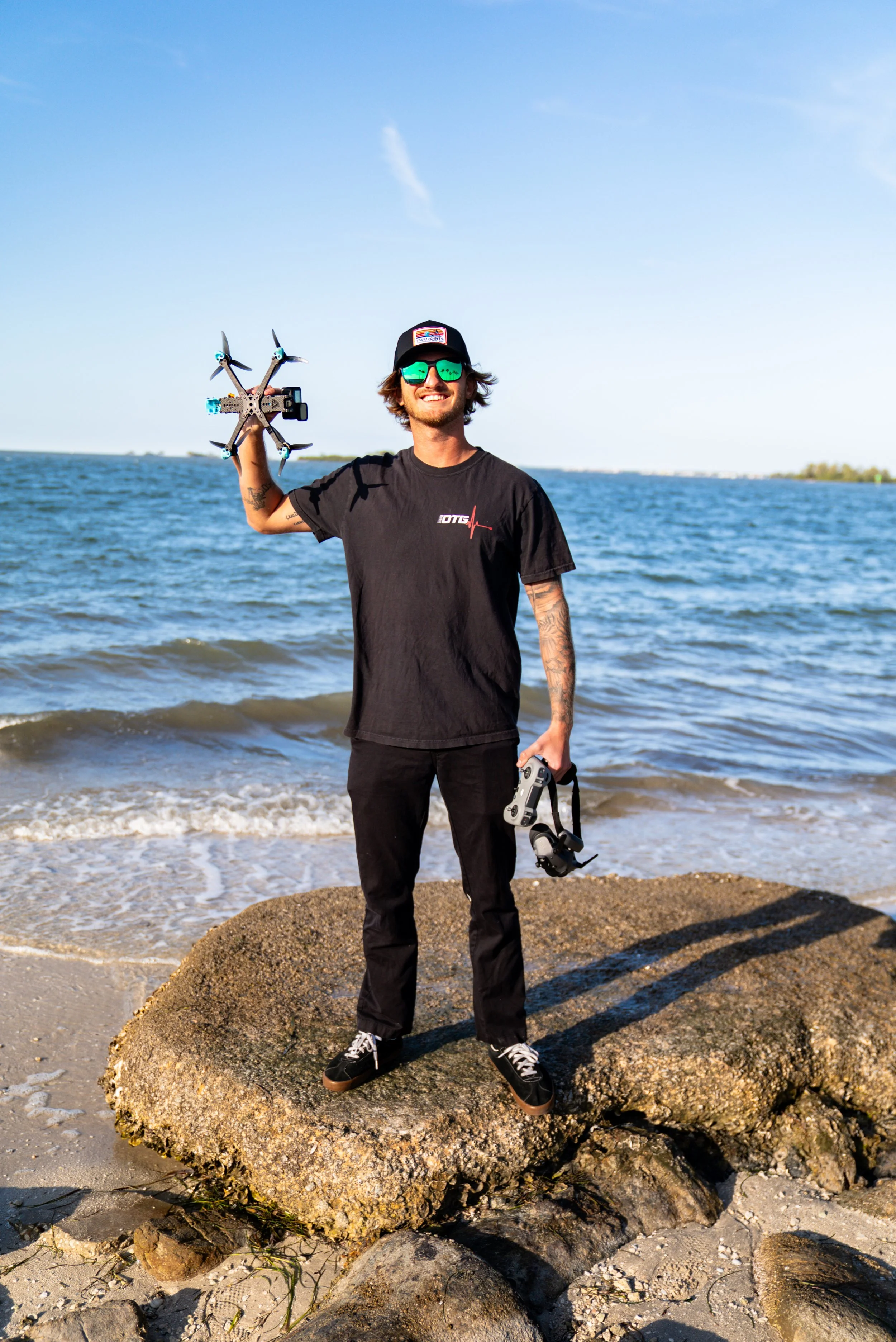 A man standing on a large rock at the beach holding a drone in his right hand and a drone controller in his left hand, wearing sunglasses, a black baseball cap, a black t-shirt, and black pants.