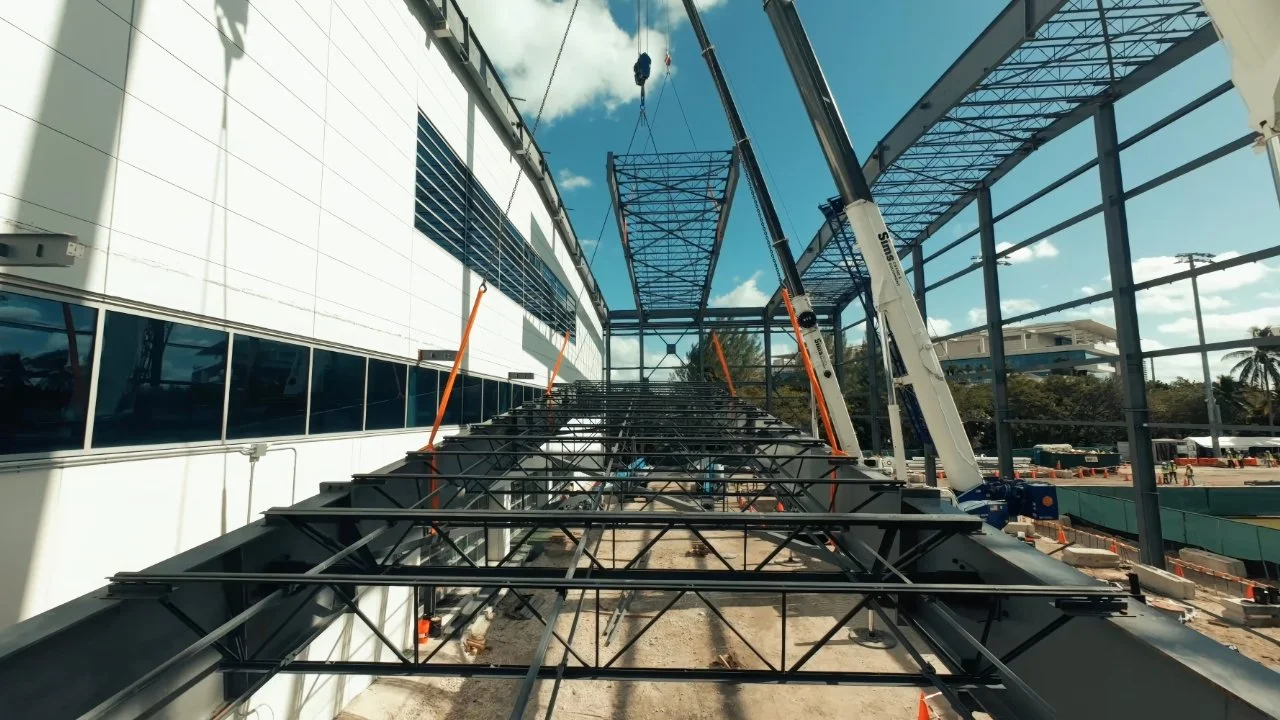Drone footage of a construction site with steel framework, a crane lifting a section, and workers in the background.