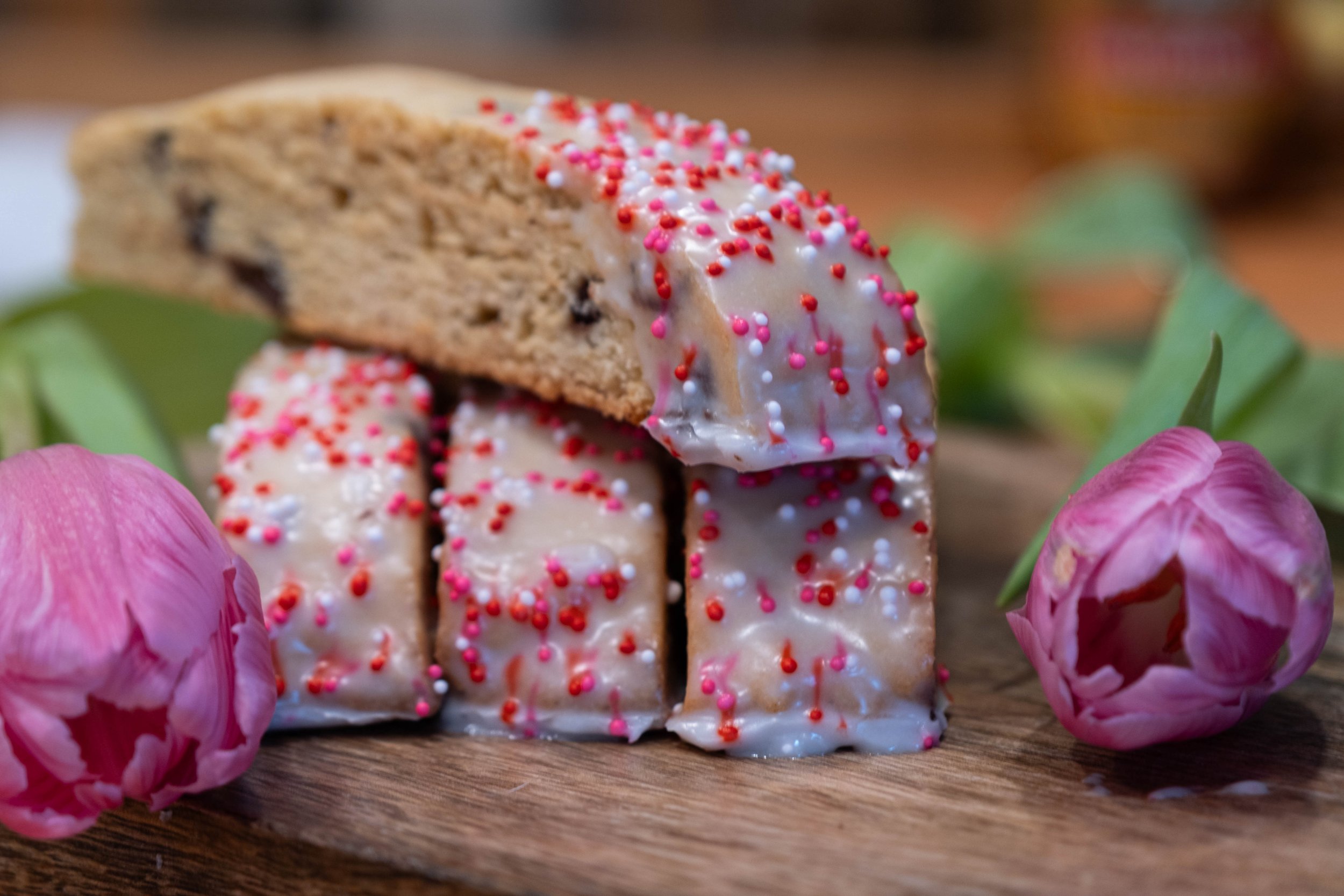 A slice biscotti sitting on top of three vanilla, chocolate chip biscotti with almond icing with pink and red sprinkles. The biscotti's are on a wooden board, surrounded by pink tulip flowers.