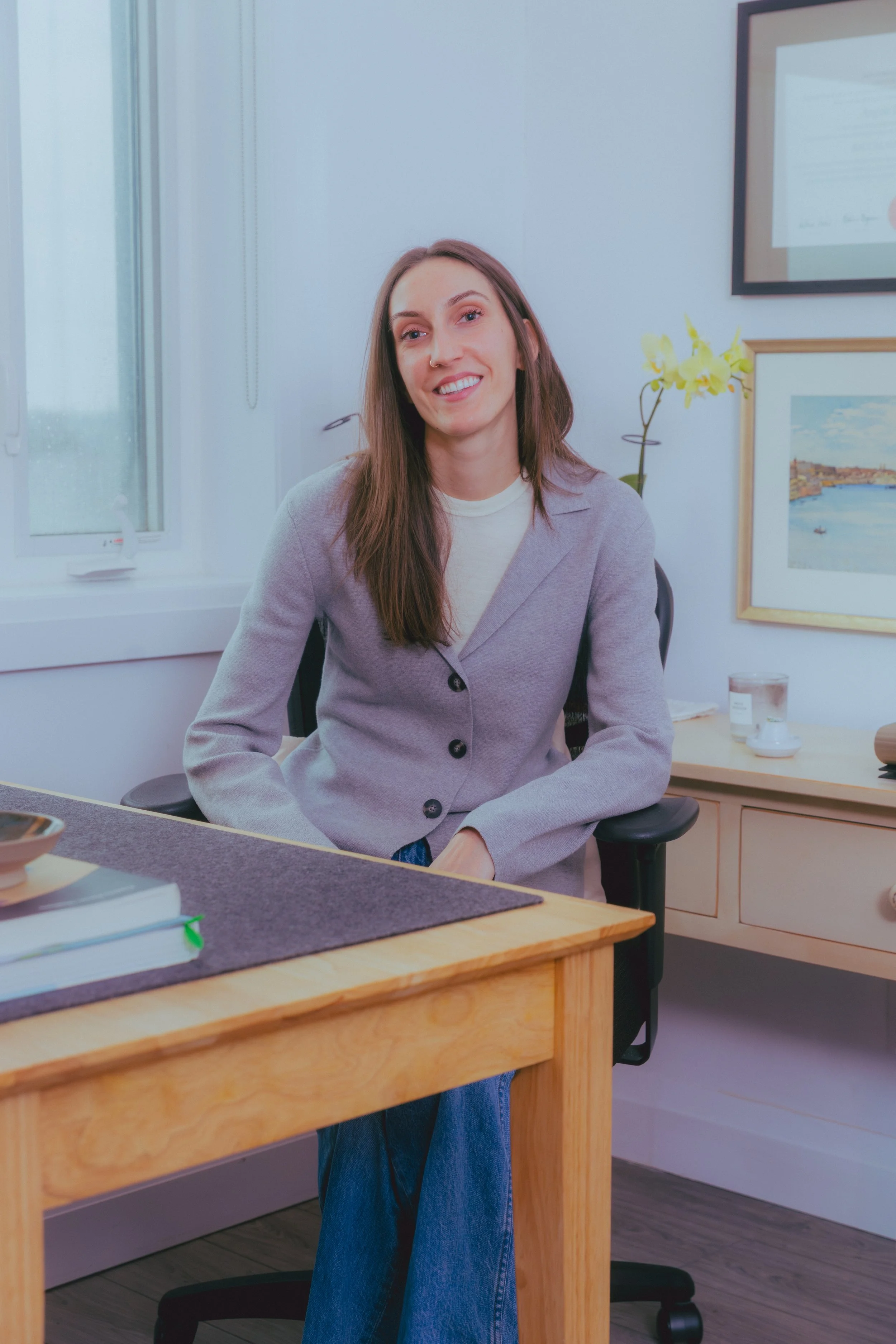 A female therapist sits smiling an office desk. A yellow orchid, framed watercolor painting, and framed university diplomas are visible in the background.