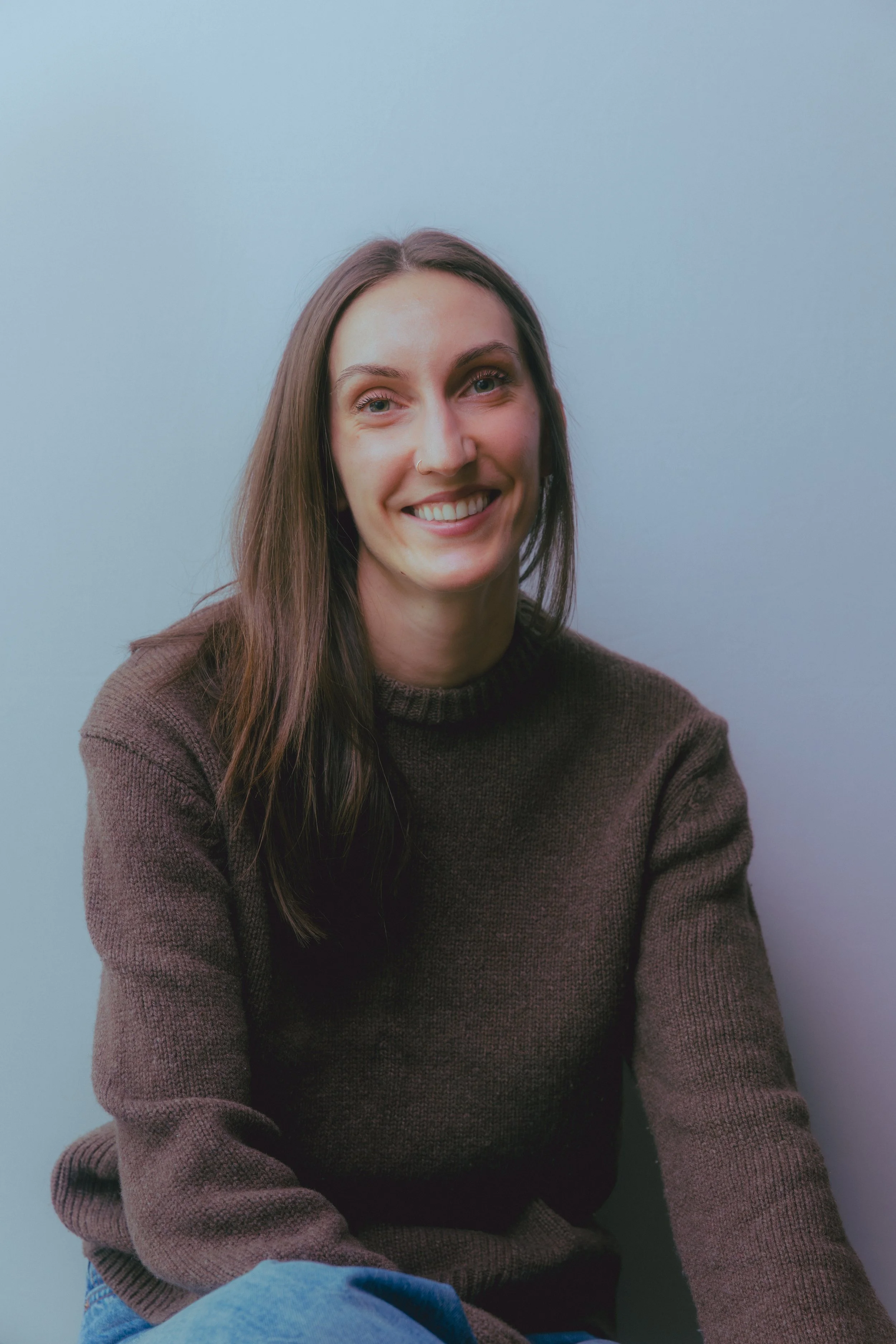 A female therapist with long brown hair wearing a brown wool sweater sits smiling against a soft white wall in a light-filled room.