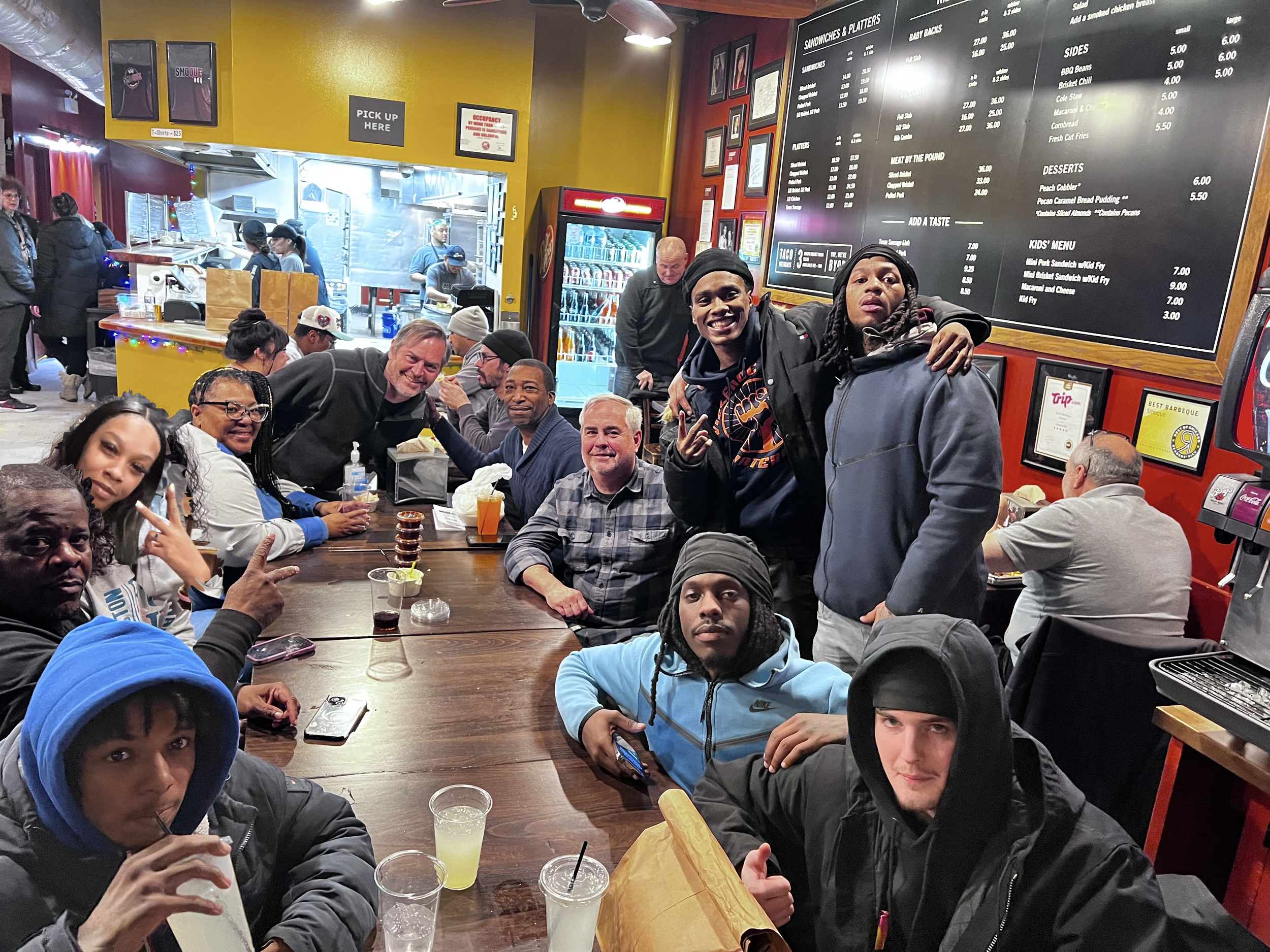 A group of diverse people enjoying a meal at a restaurant with a large menu on the wall, some holding drinks, with the kitchen visible in the background.