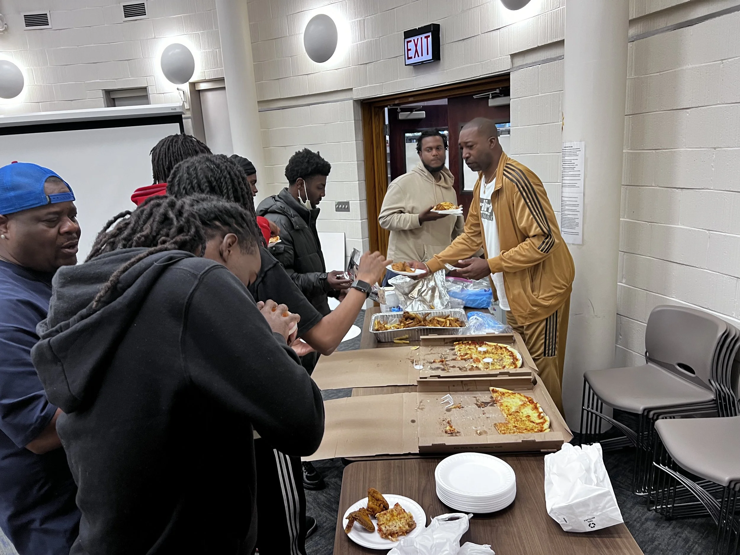Group of people in line at a pizza and buffet food table, with plates and pizza boxes on the table, inside a room with white walls and exit sign.