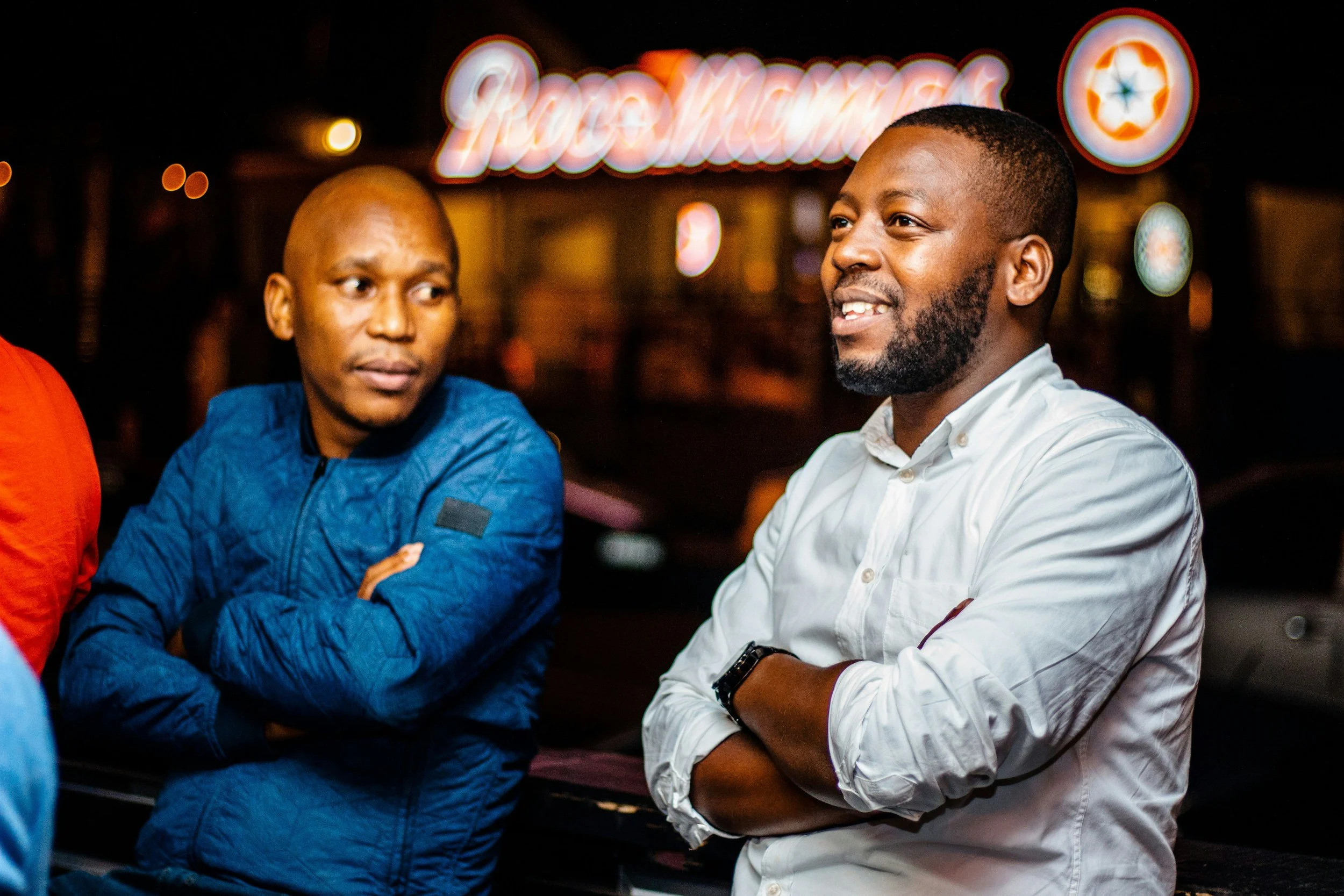 Two men standing and talking outside at night under bright lights with illuminated sign in the background.