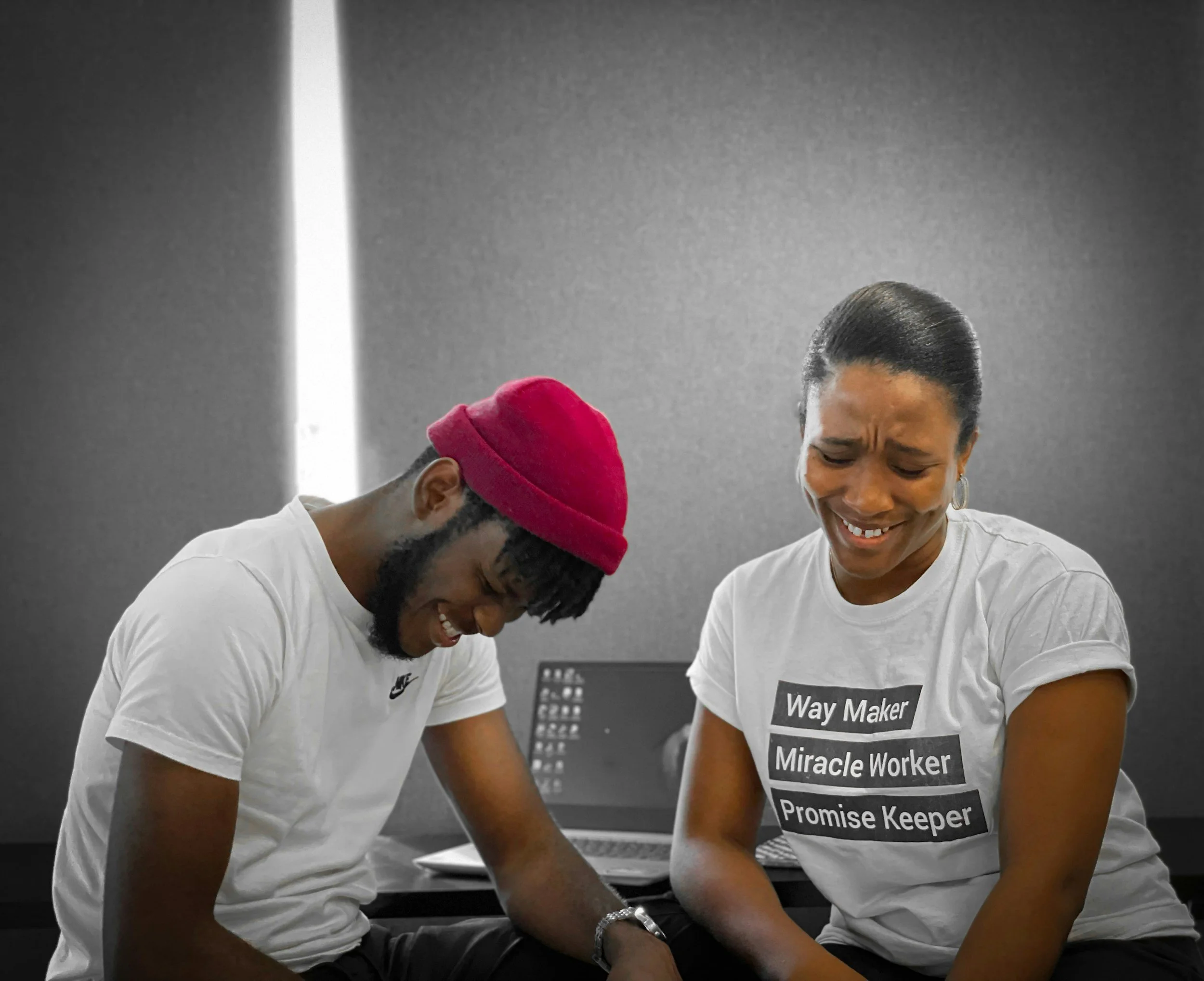 Two people sitting together, smiling and looking down, in a room with a gray wall. The man on the left wears a red beanie and a white Nike t-shirt, and the woman on the right wears a white t-shirt with motivational words printed on it.