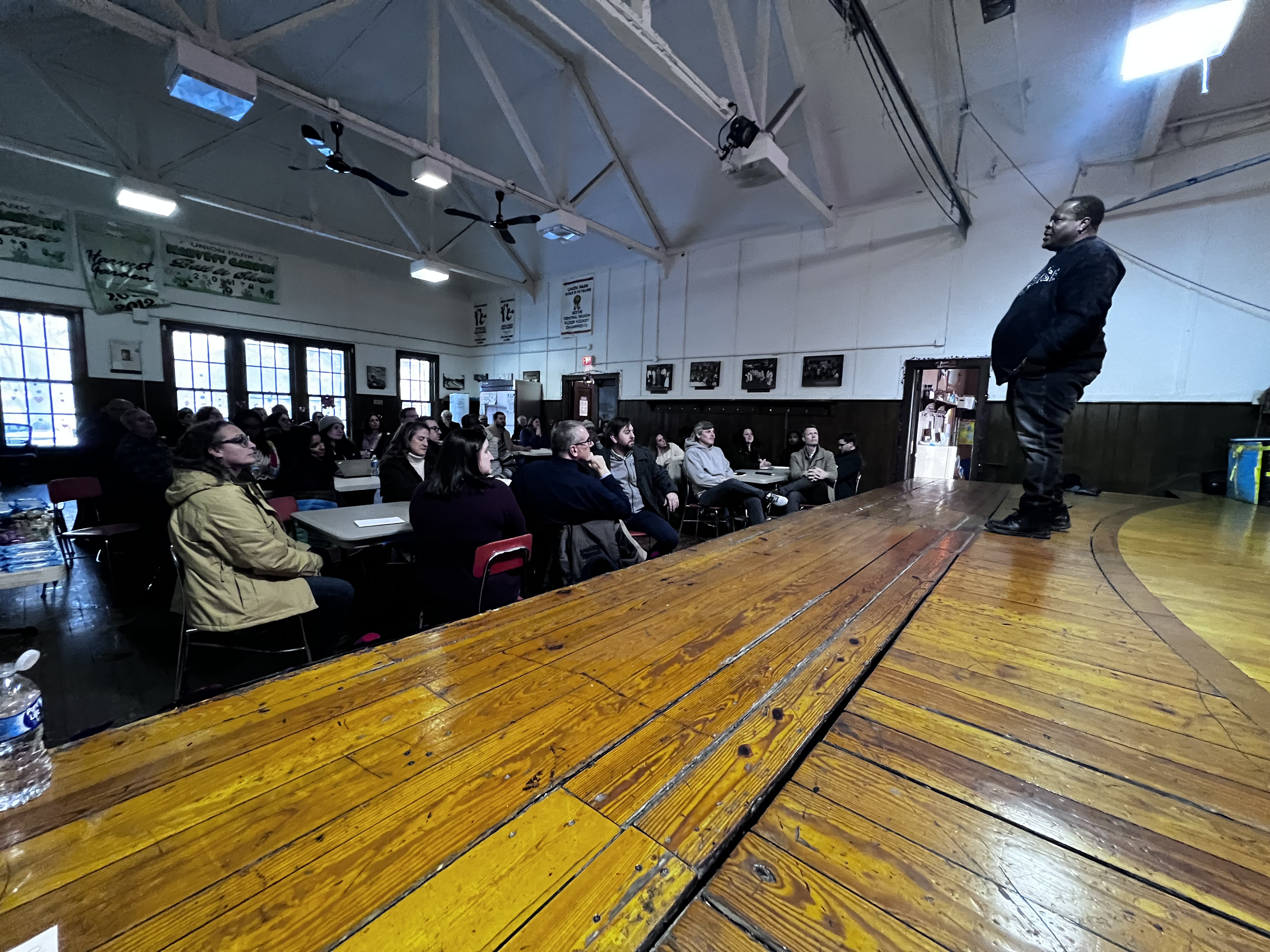 A man standing on a wooden stage addressing an audience inside a community hall or gymnasium. The audience is seated at tables, listening attentively. The room features windows, ceiling fans, and hanging banners.