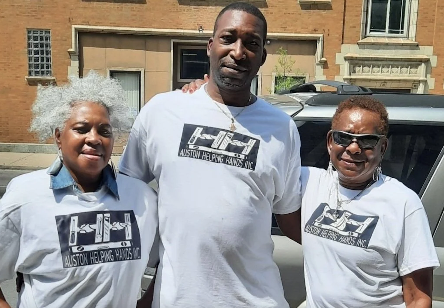 Three smiling people standing outdoors in front of a building and a vehicle, wearing matching white T-shirts with a logo that reads "Auston Helping Hands Inc."