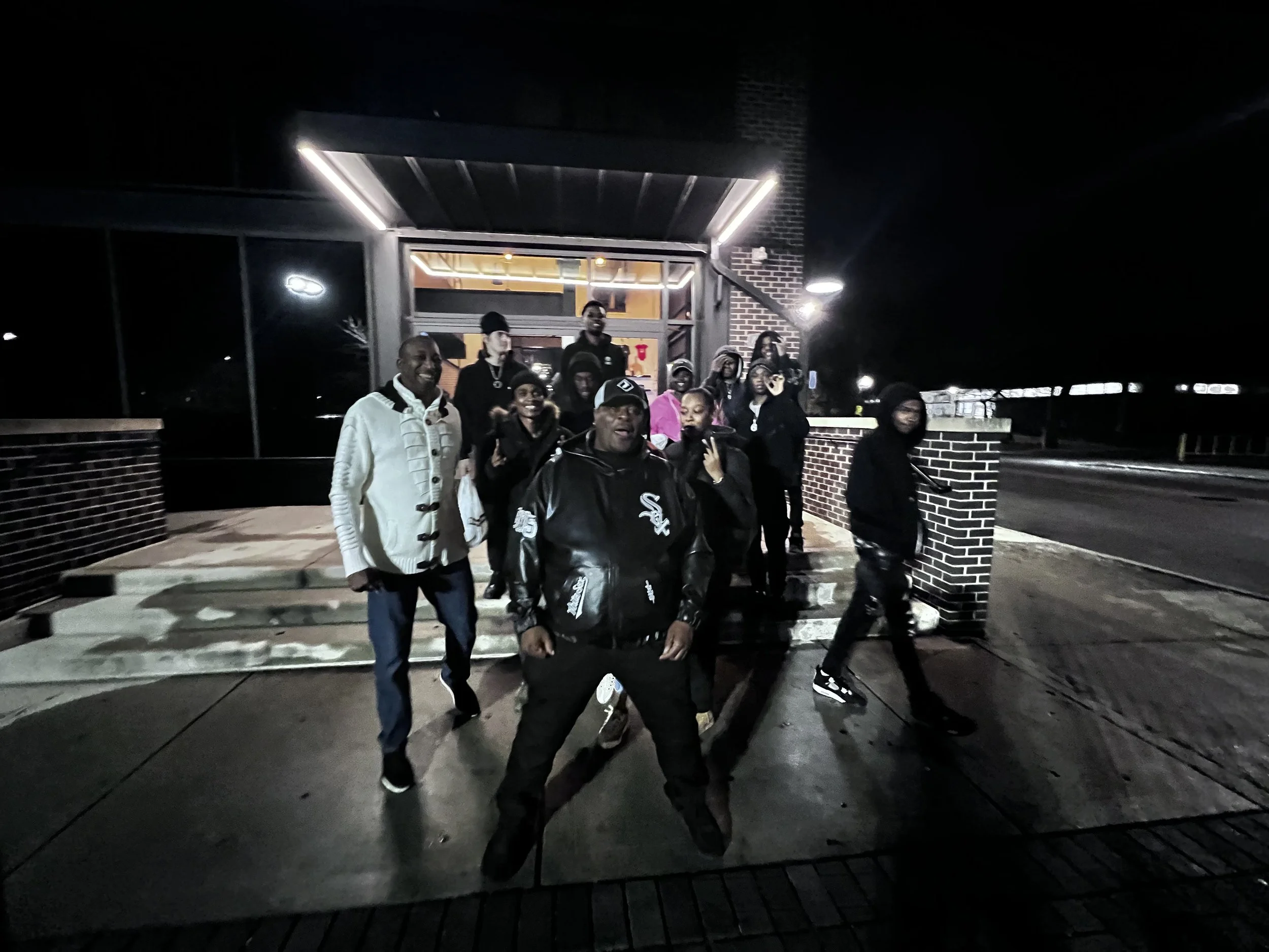 A group of people standing outside at night on a sidewalk in front of a building with brick walls and large windows.