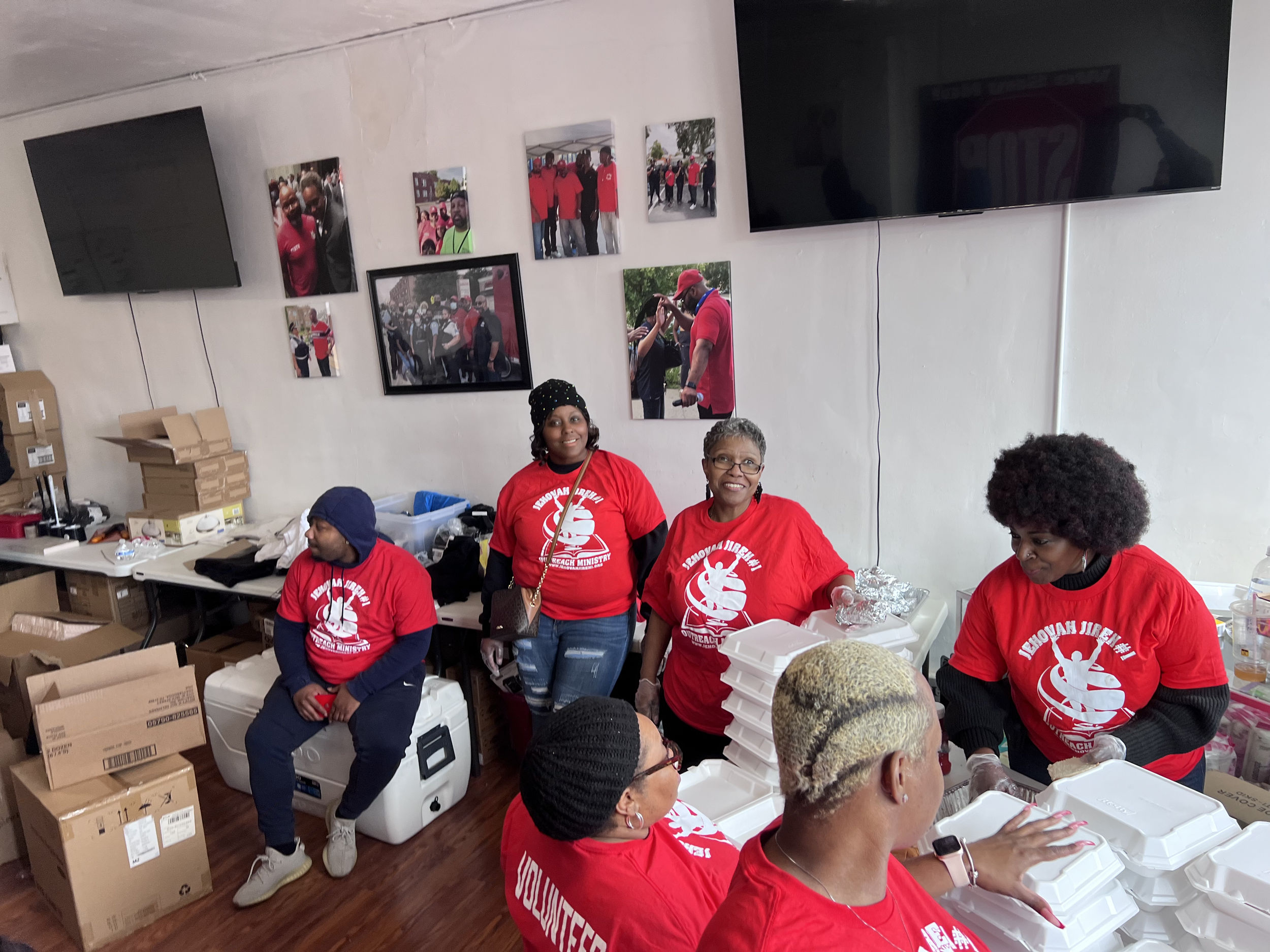 Group of volunteers in red shirts helping pack food in a room with photos and televisions on the wall.