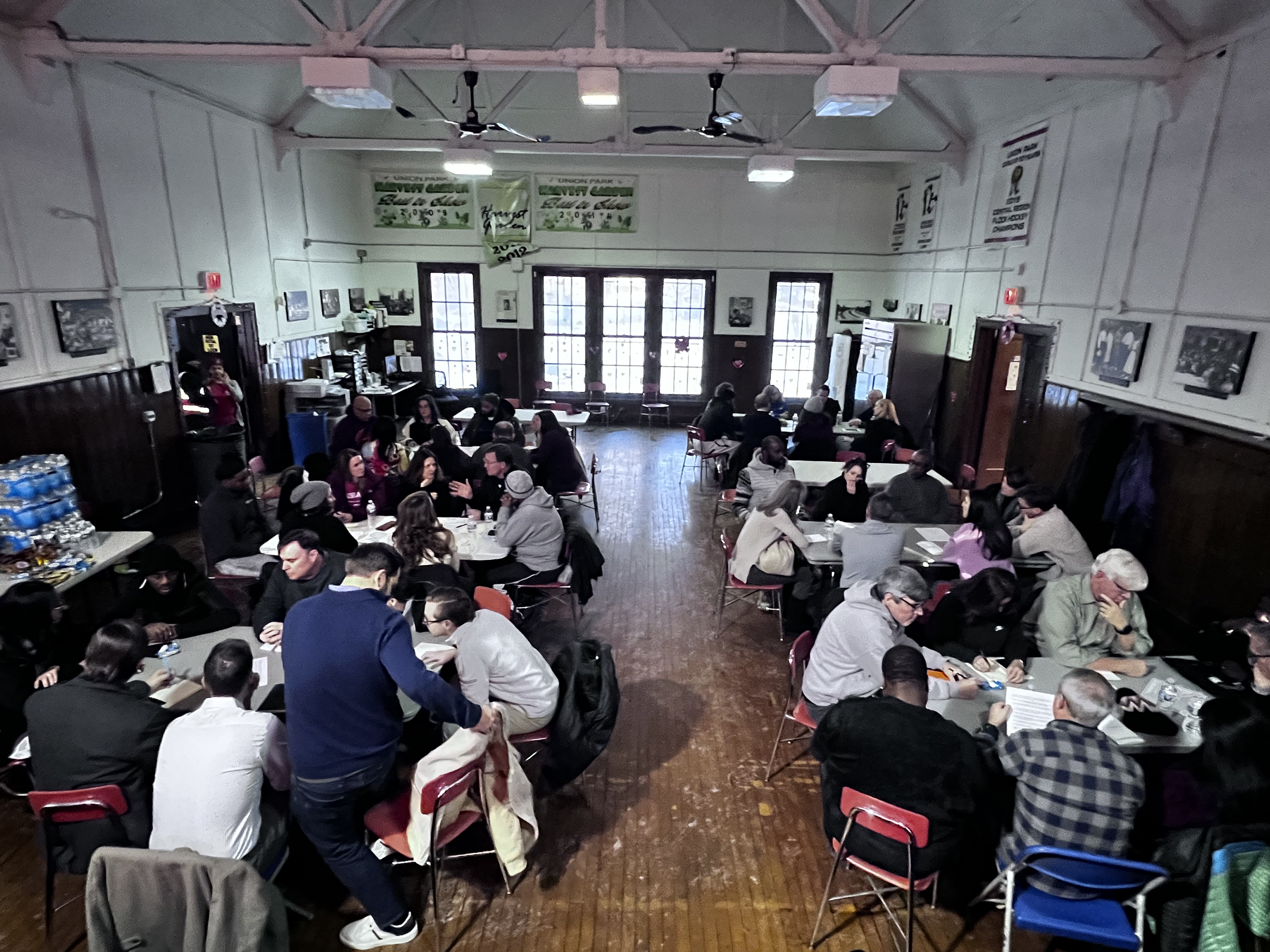A large room filled with people sitting at tables, engaging in conversations, with some standing. The room has wooden floors, high ceilings, ceiling fans, and large windows at the back. Walls are decorated with posters and framed pictures, and there are coats hanging on hooks along the right wall.