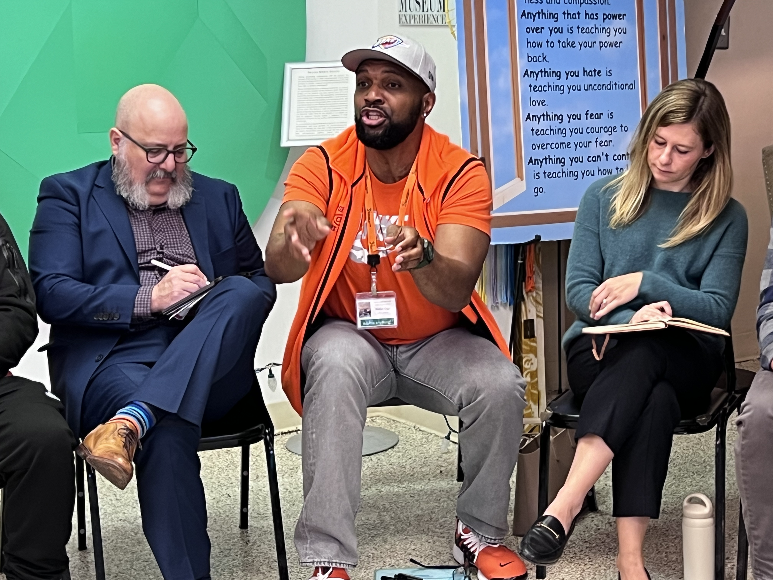 A group of three people sitting in chairs engaged in a discussion, with a man in the middle speaking and gesturing with his hands.