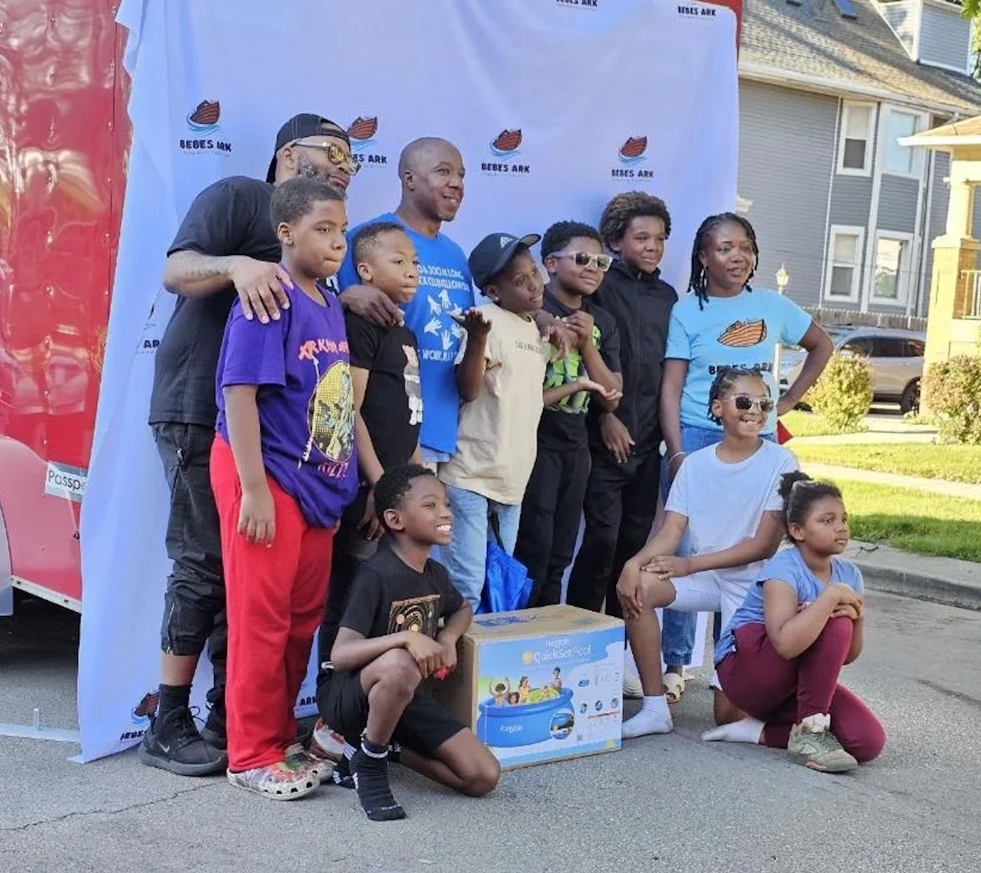 A group of children and adults posing outdoors in front of a backdrop with the logo 'Bebes Ark.' Some children are sitting or kneeling, and others are standing, with one holding a boxed toy. The people are smiling for a photo during a community event.